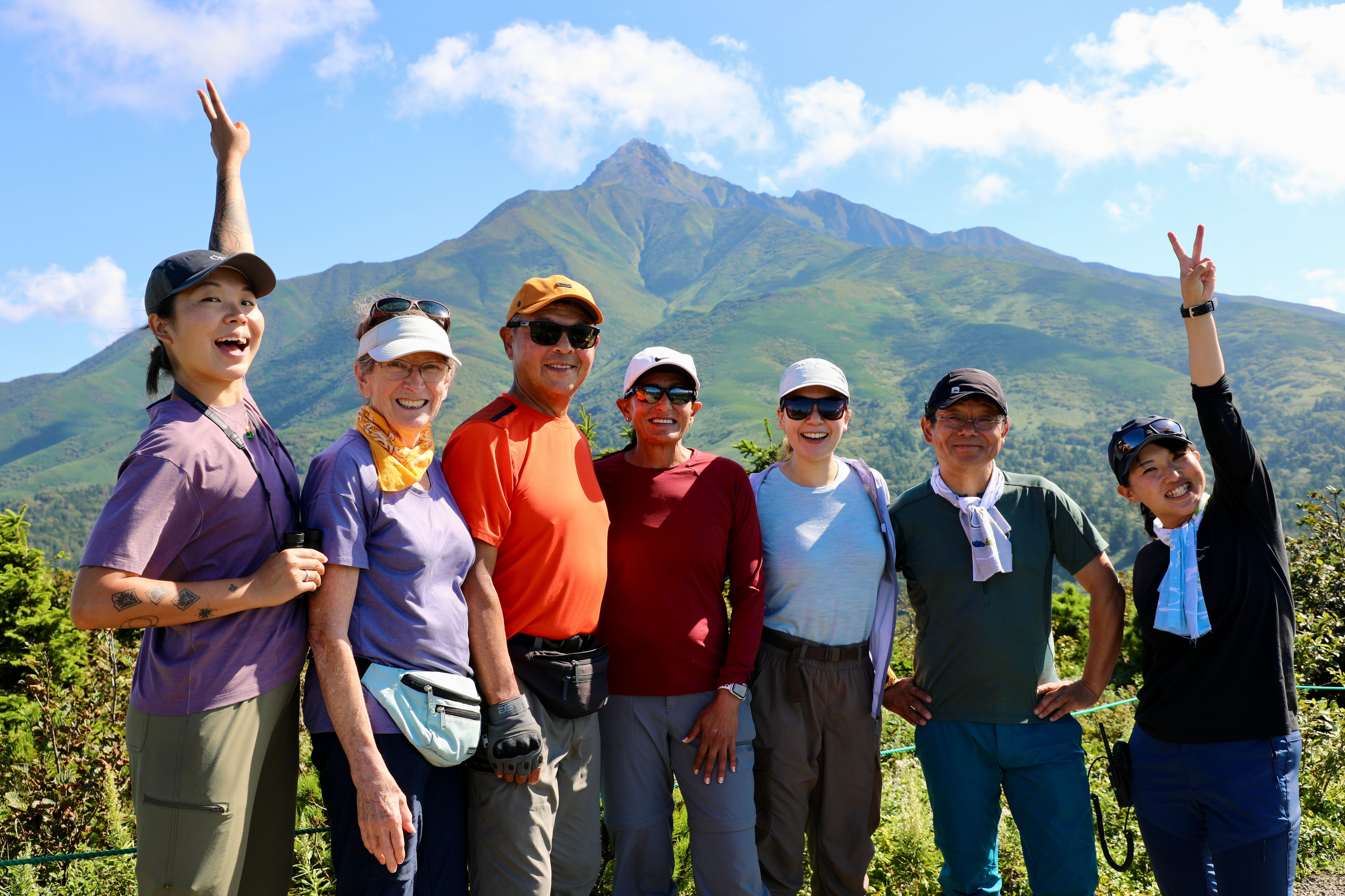 A group of hikers pose in front of Mt. Rishiri on Rishiri Island, Hokkaido. Some are putting their fingers in the air in a kind of "peace" sign. Photo credit: Samir Patel