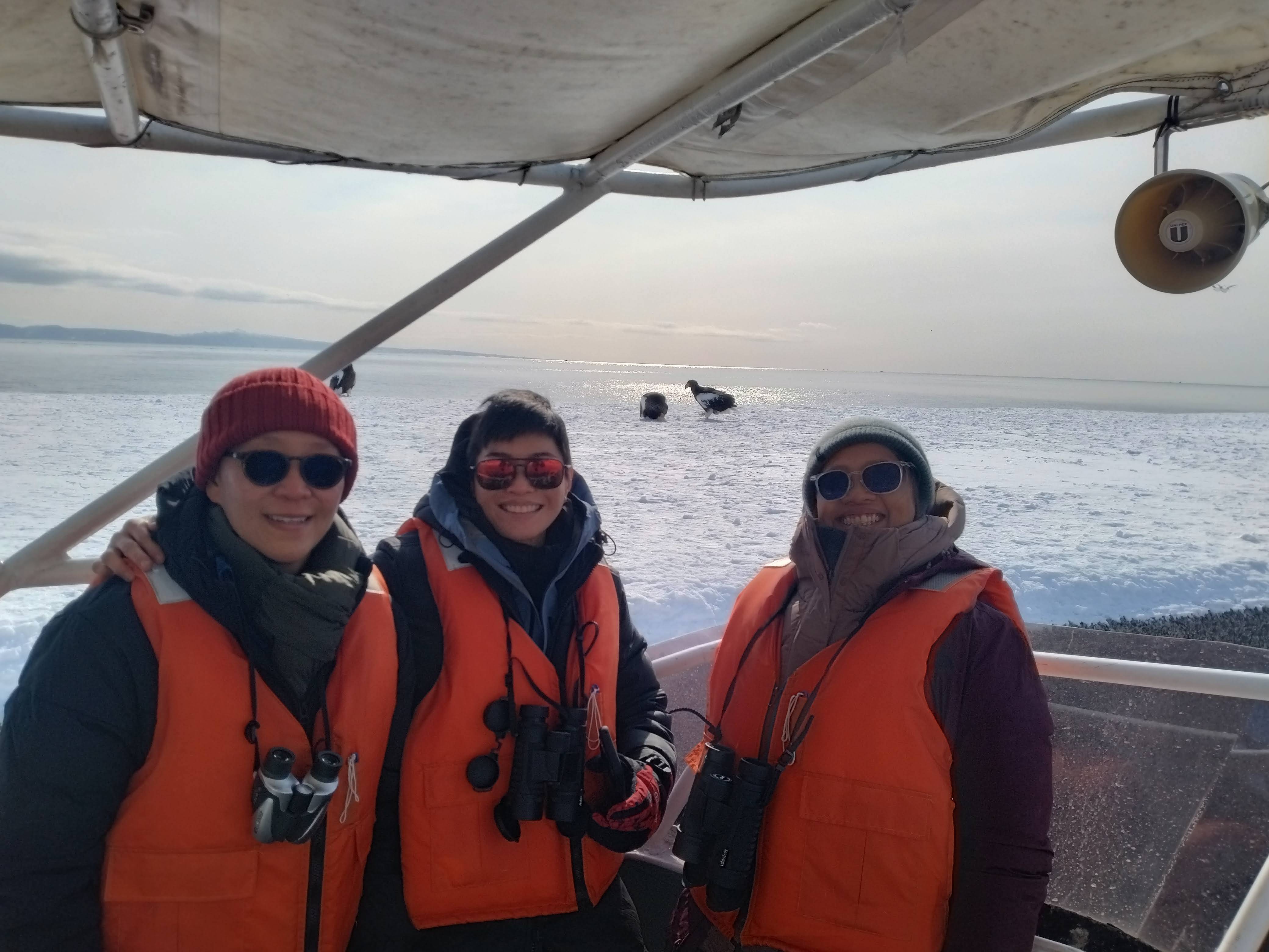 A group of three people on a cruise boat smile at the camera. They are all wearing life jackets, suggesting the boat is on the water. Behind them are two Steller's sea eagles.