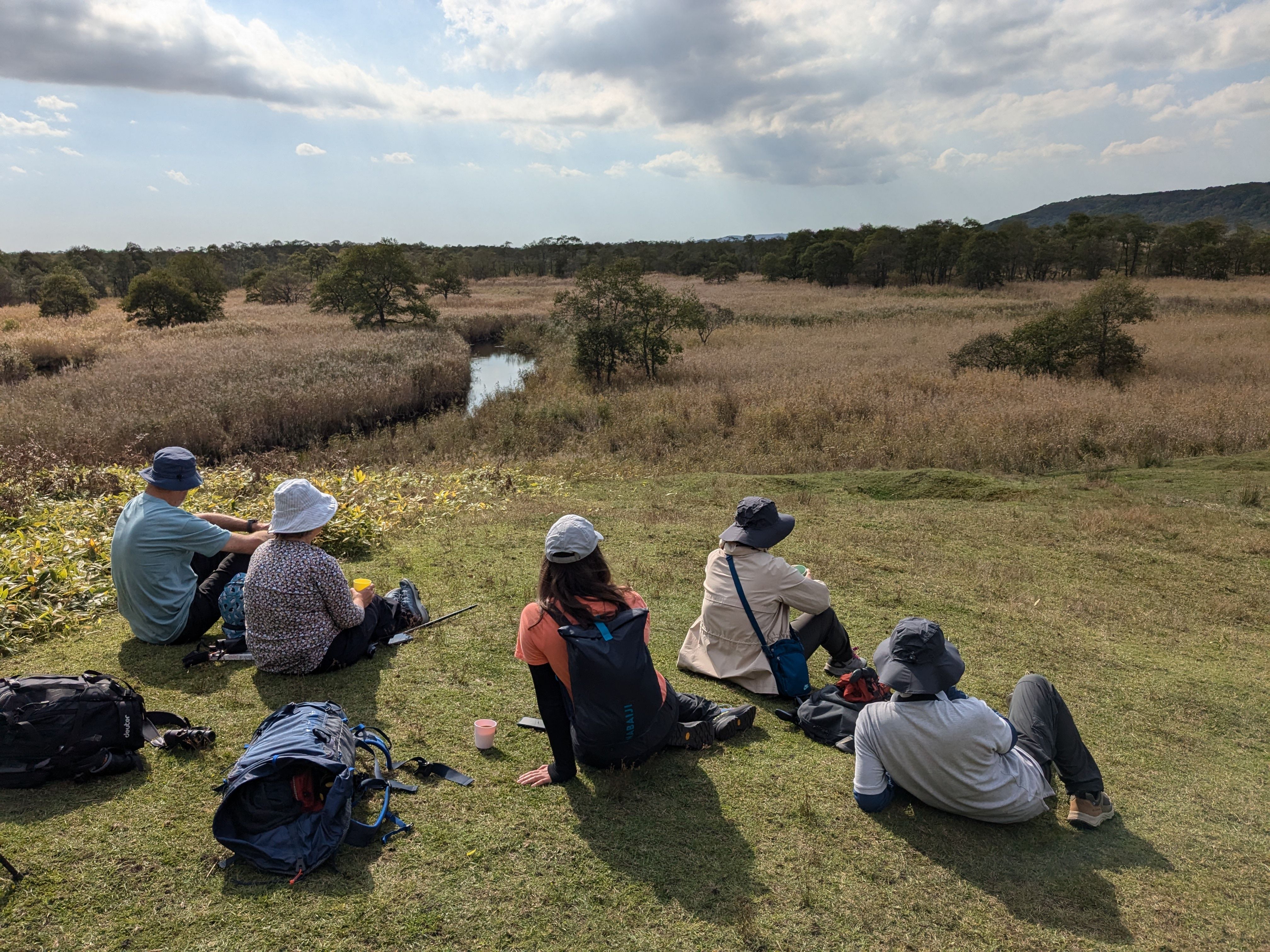 A group of walkers sit looking out over the Kushiro Wetlands from atop a hill. It's a beautifully sunny day and a river winds its way through the wetlands below.