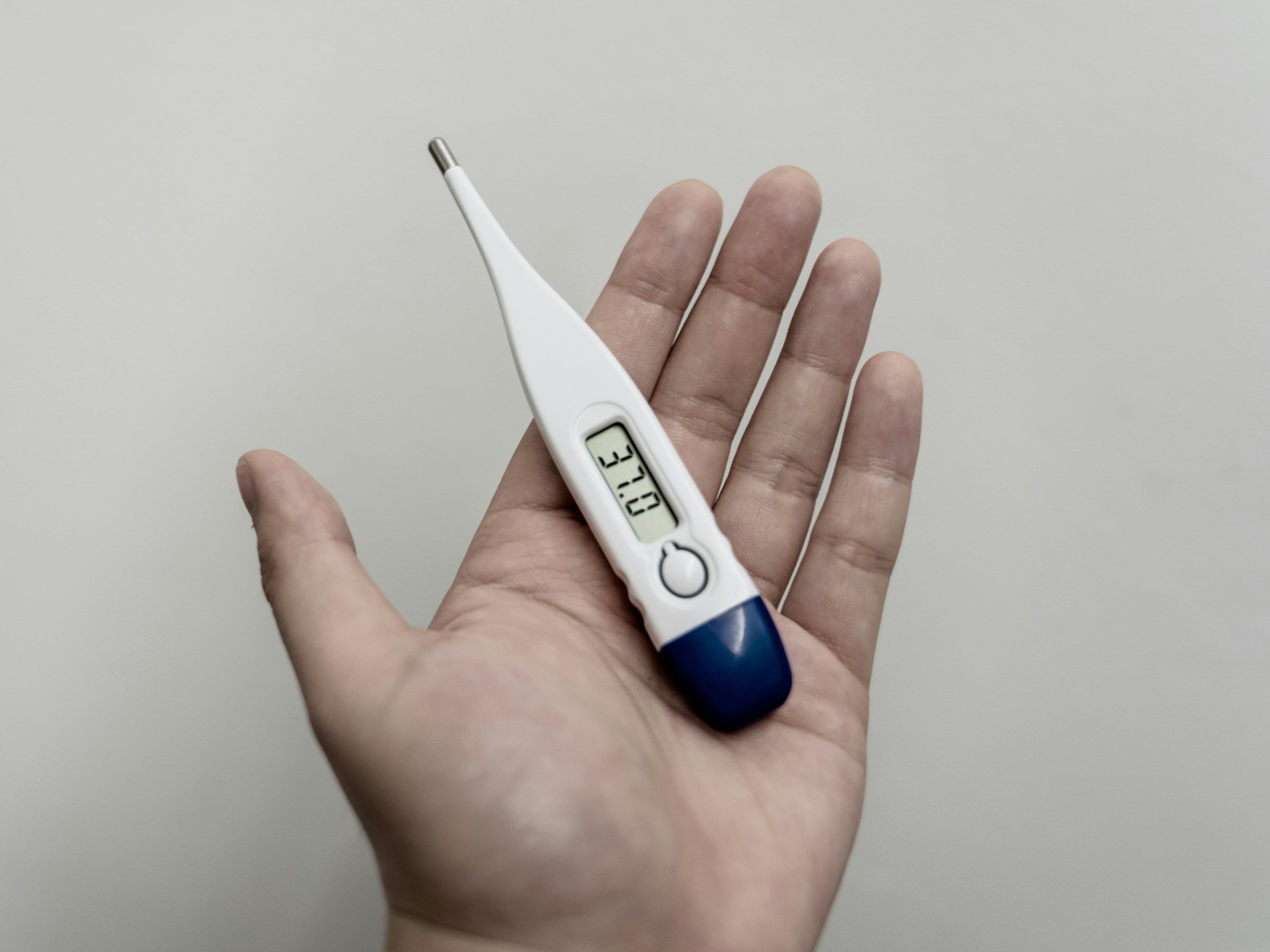 A person's open hand holds a digital thermometer that displays a body temperature reading of 37.0 degrees Celsius.