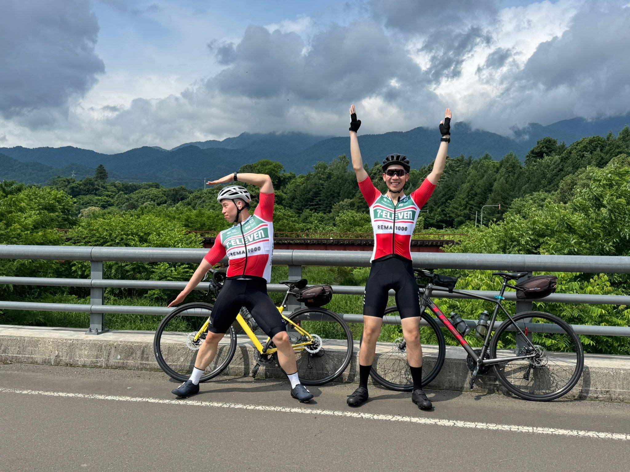 Two cyclists pose in their cycling gear on a bridge, their bikes behind them. They are wearing "7 Eleven" cycling gear and are making the numbers "7" and "11" with their arms.