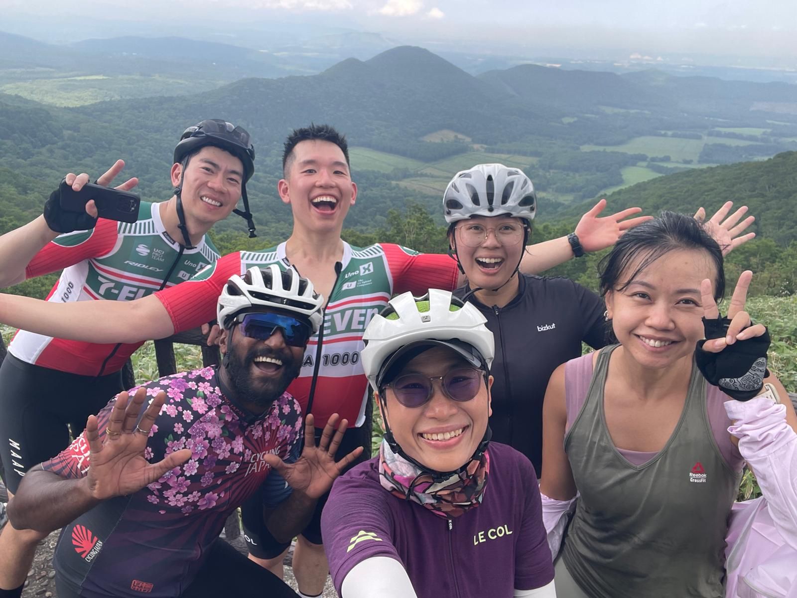 A group of cyclists take a selfie in front of the view of Karikachi Pass. The scenery of the Tokachi Plains is clearly visible behind them.
