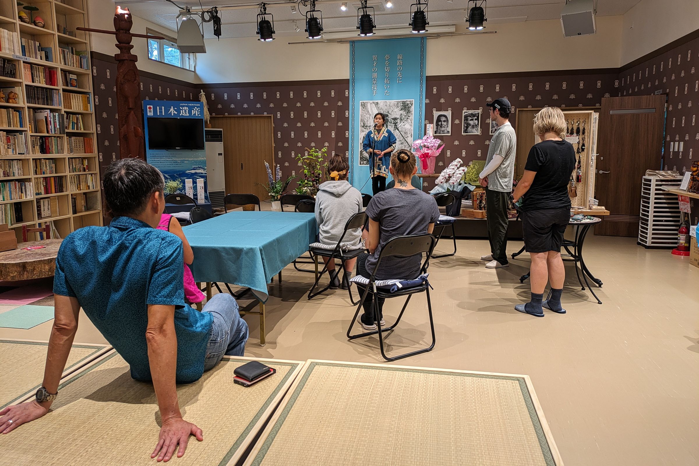 Guests sit in a room while an Ainu woman gives a presentation on a stage. She is dressed in traditional Ainu clothing.
