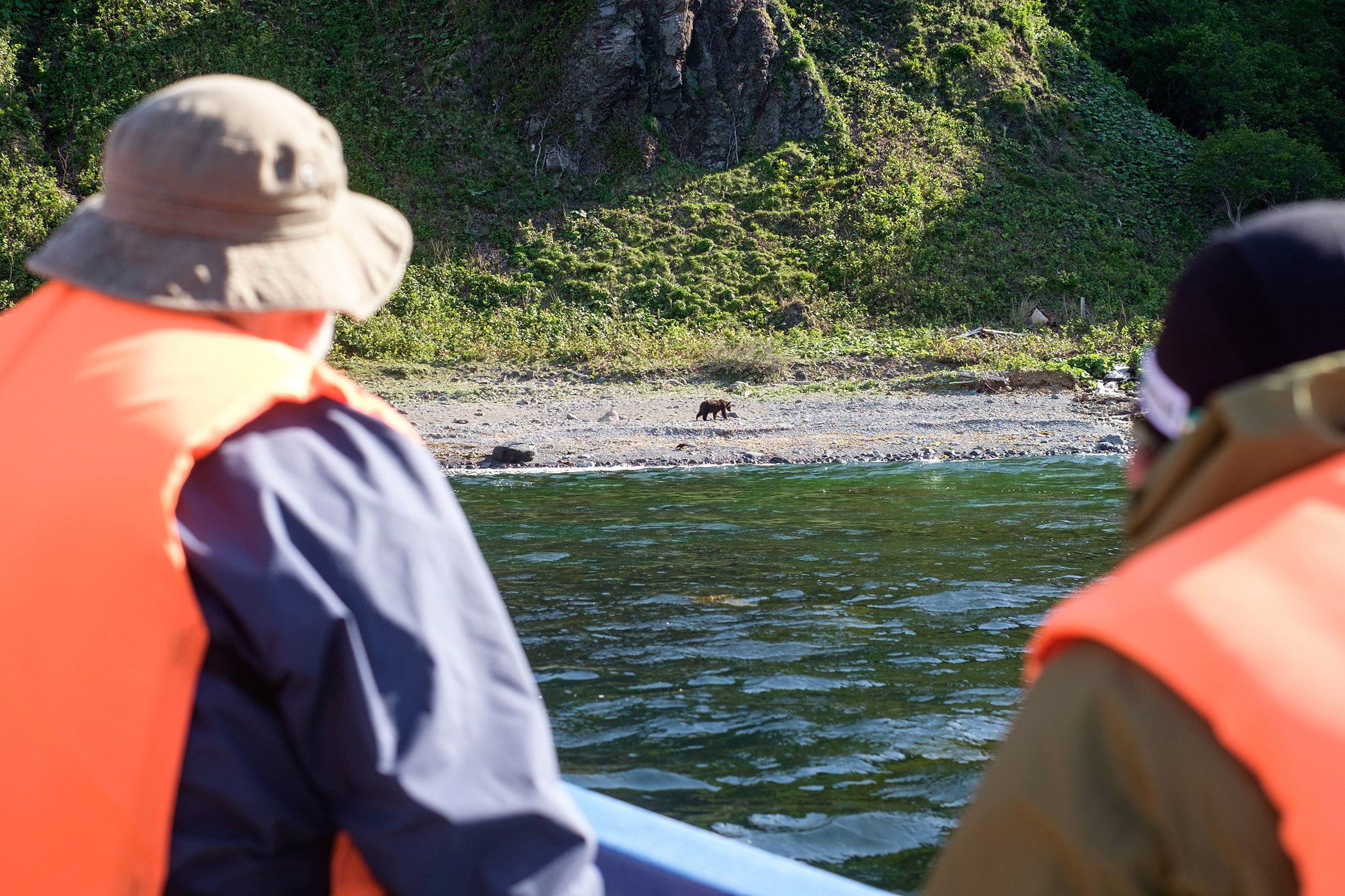 Two wildlife watchers wearing orange life jackets and riding a small boat look at a Brown Bear walking along the shore