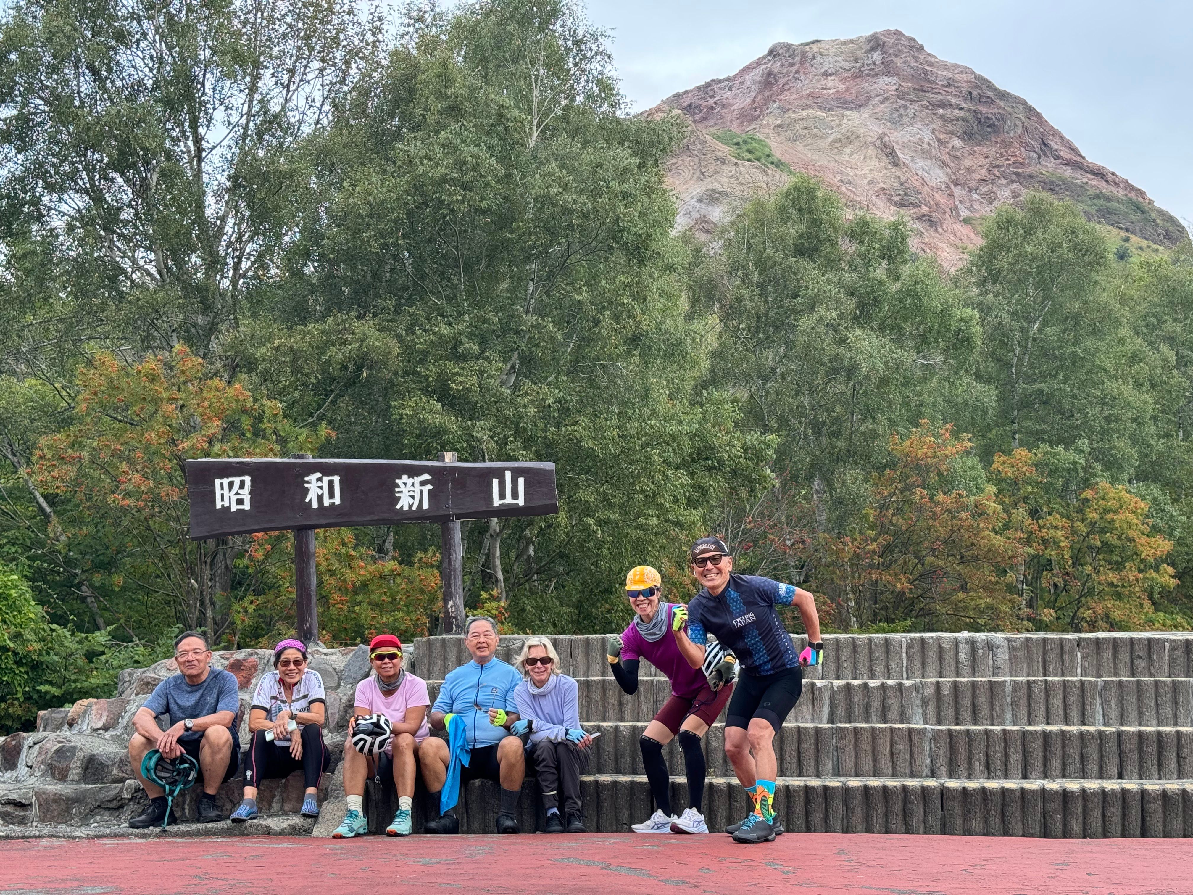 A group of seven cyclists smile at the camera in front of Mt. Showa-Shinzan, an active volcano in Hokkaido. In the background behind them, the volcano's lava dome is visible.