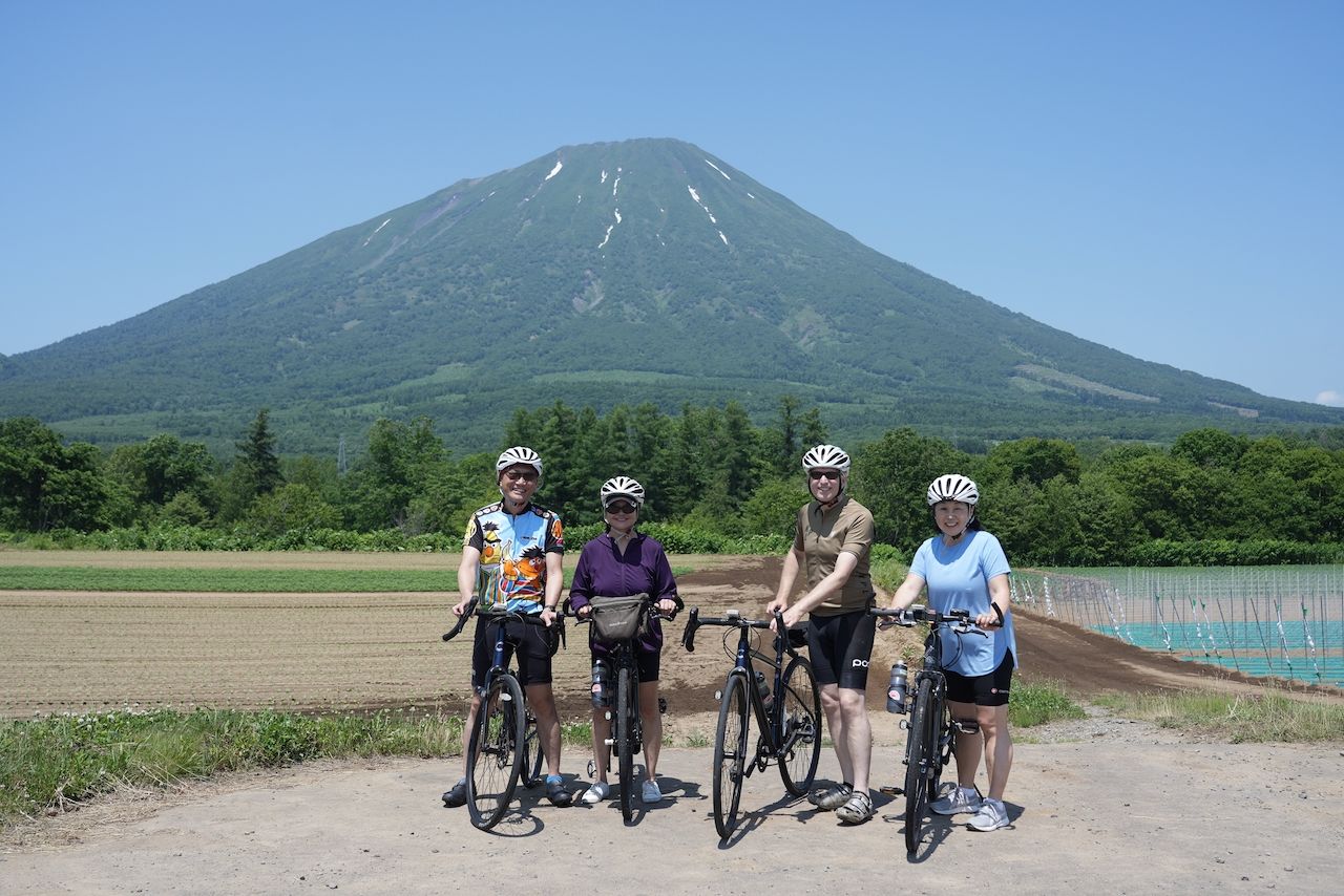 Four cyclists, two couples, stand with their bikes in front of majestic Mt. Yotei under a clear blue summer sky. Forests and farmland stretch between the cyclists and the mountain.