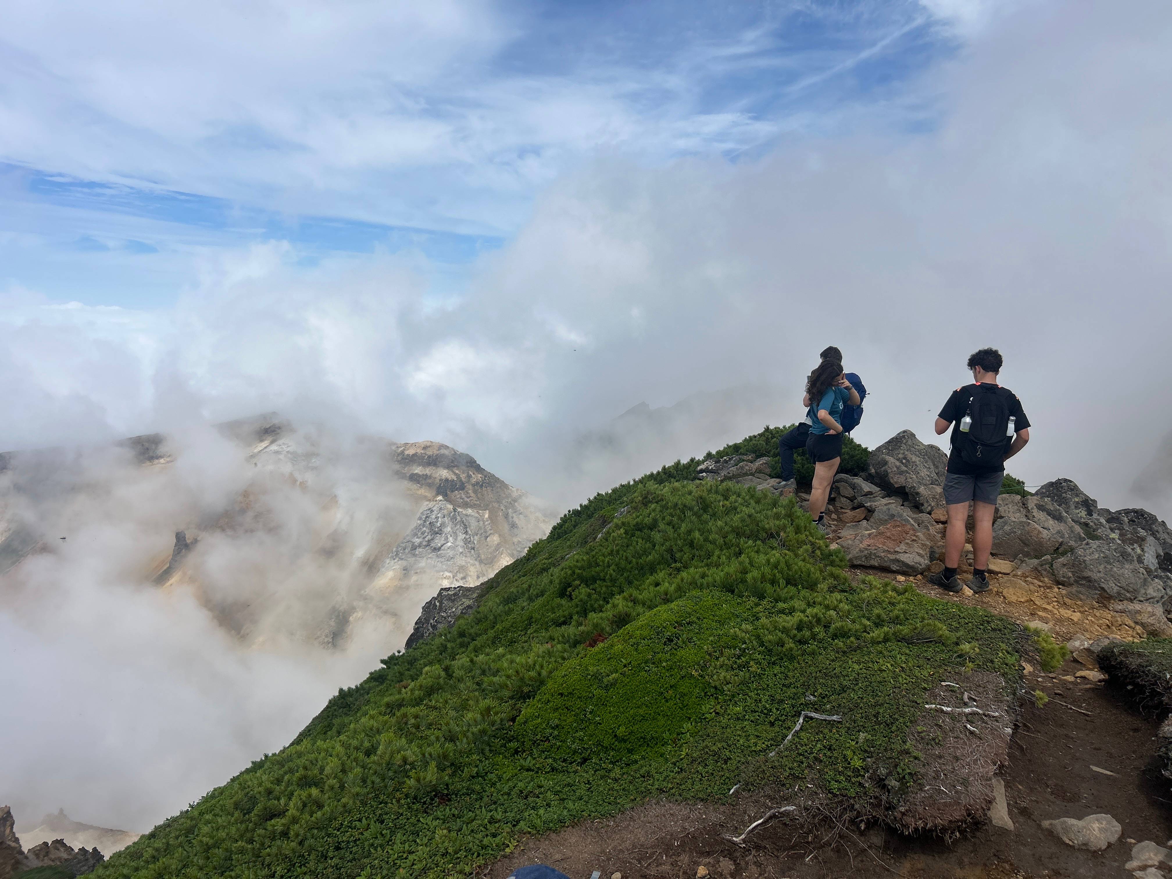 Two hikers stand atop a Rocky Mountain ridge, peering into steam rising from a valley beyond. A volcanic crater is just visible through the steam.