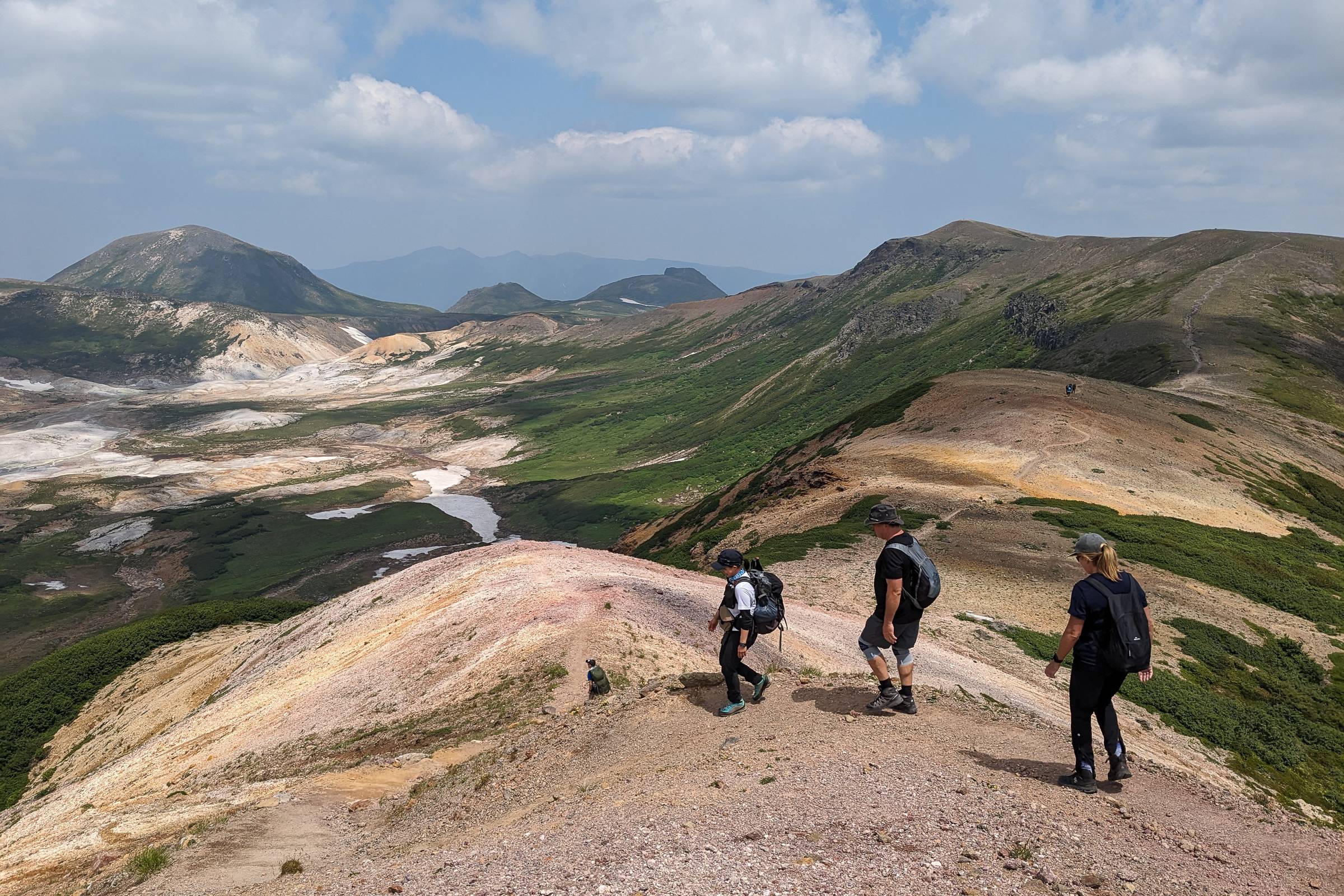 A group of hikers descent down the ridgeline on Mt. Asahidake. There are many mountains in the distance and the weather is fine.