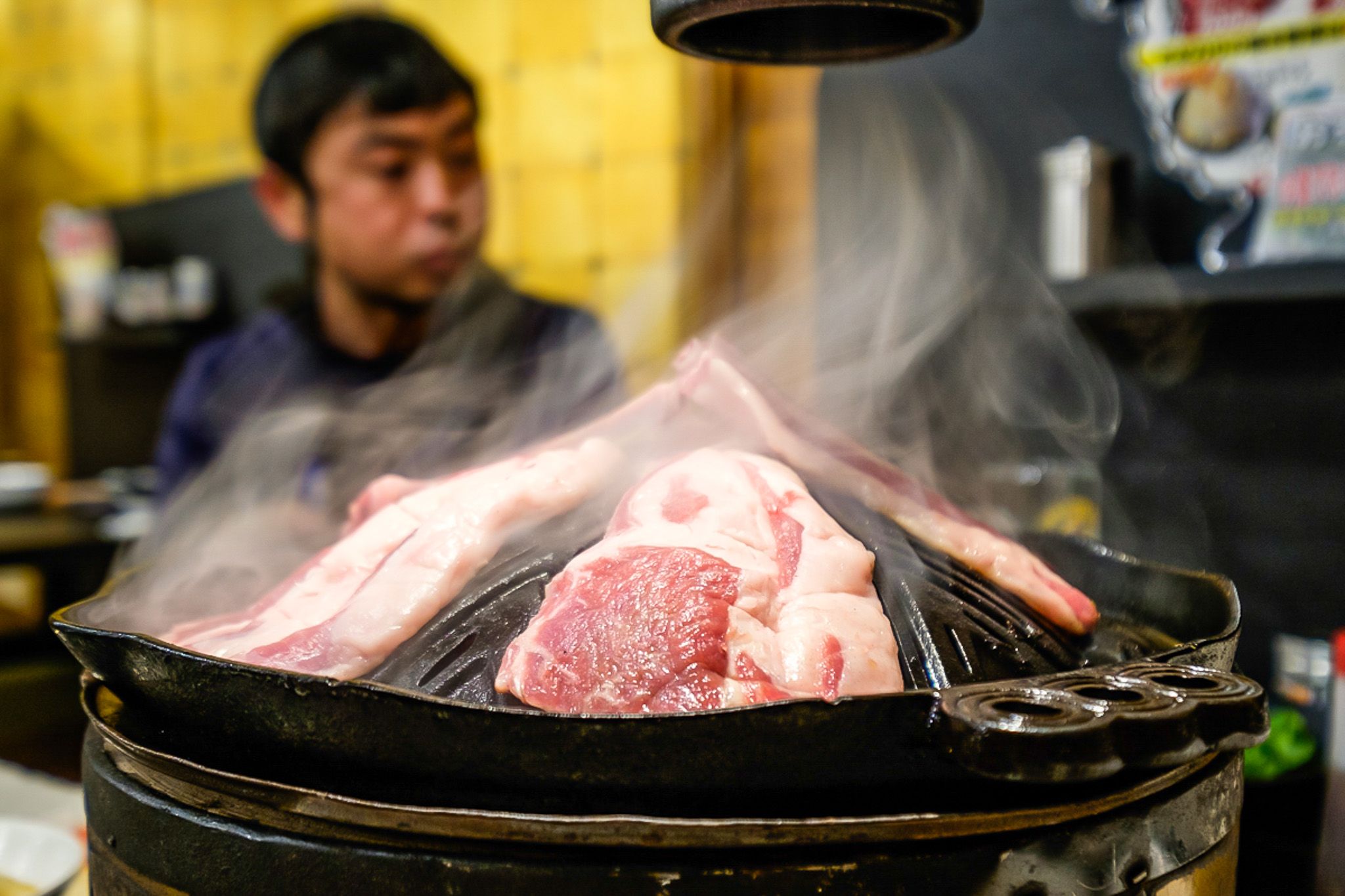 Slices of lamb sit on a black iron grill, smoke rising from them as they cook. The grill is a conical shape with grooves to carve the fat.