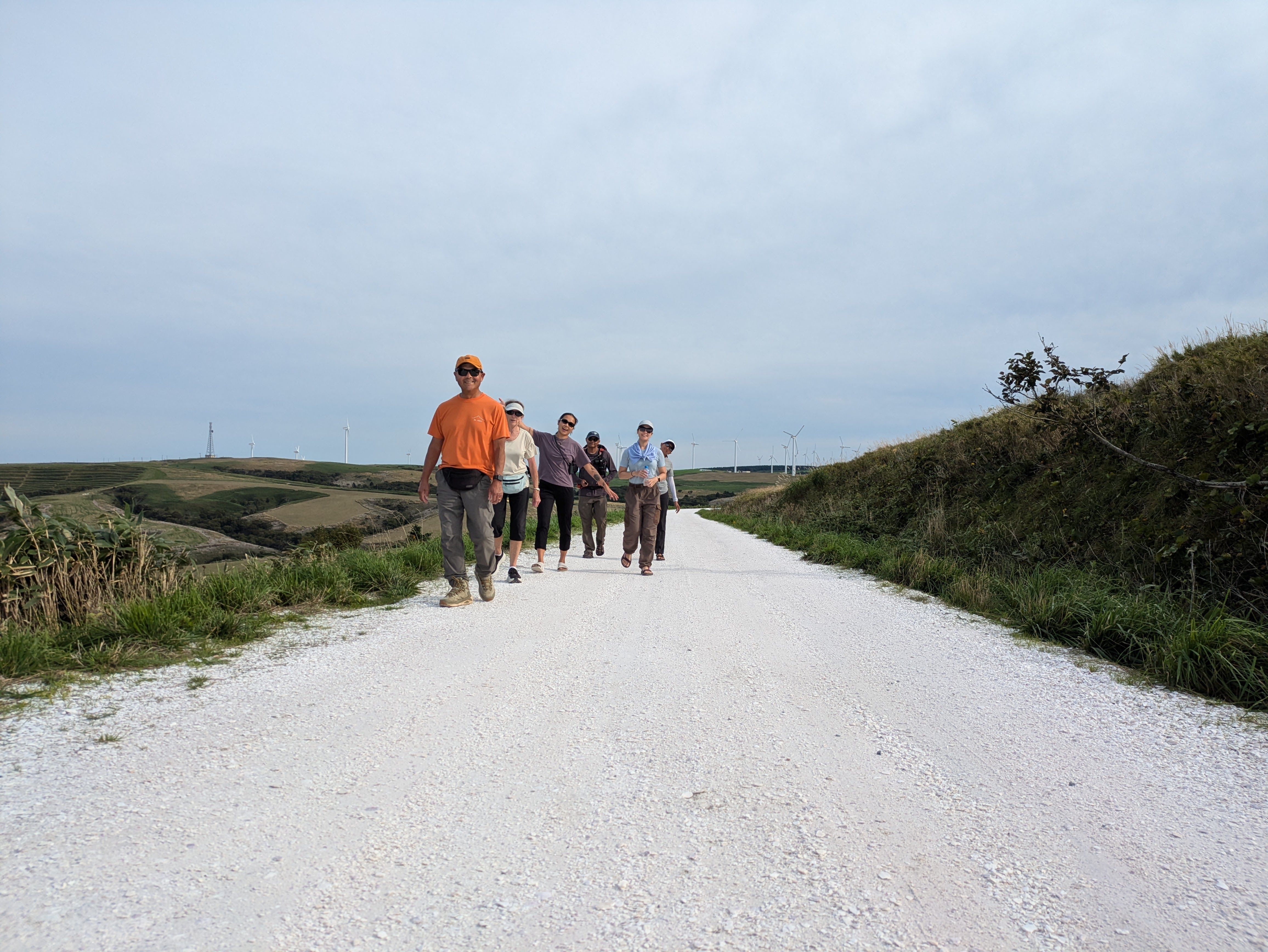 A group of walkers cheerfully walk along the White Shell Path in Wakkanai, Hokkaido. They are all smiling at the camera. It is an overcast day but the sun is trying to break through. In the distance behind the group, large wind turbines stick up out of the hills.