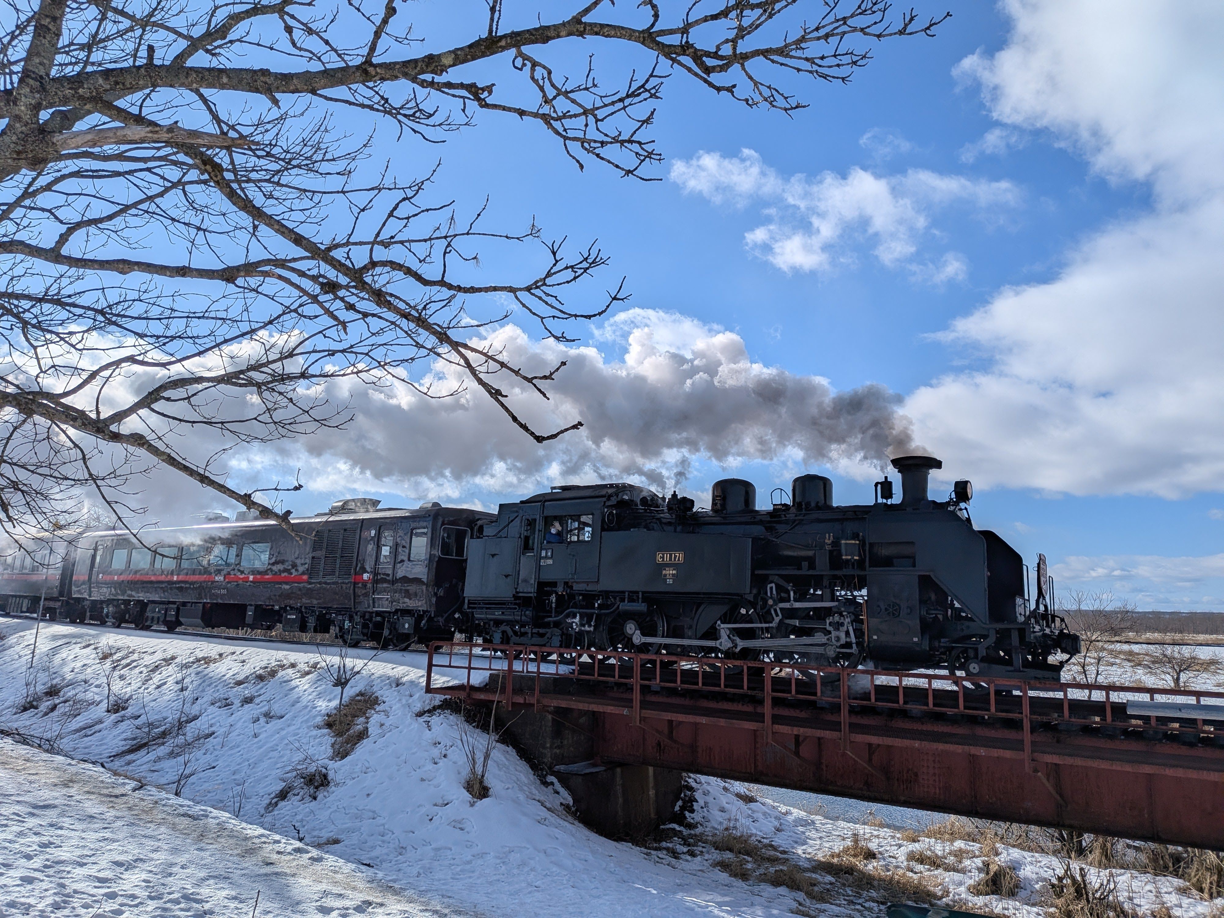 The steam train Kushiro Shitsugen SL crosses a railway bridge. Steam billows from its smokestack. It is a clear day.