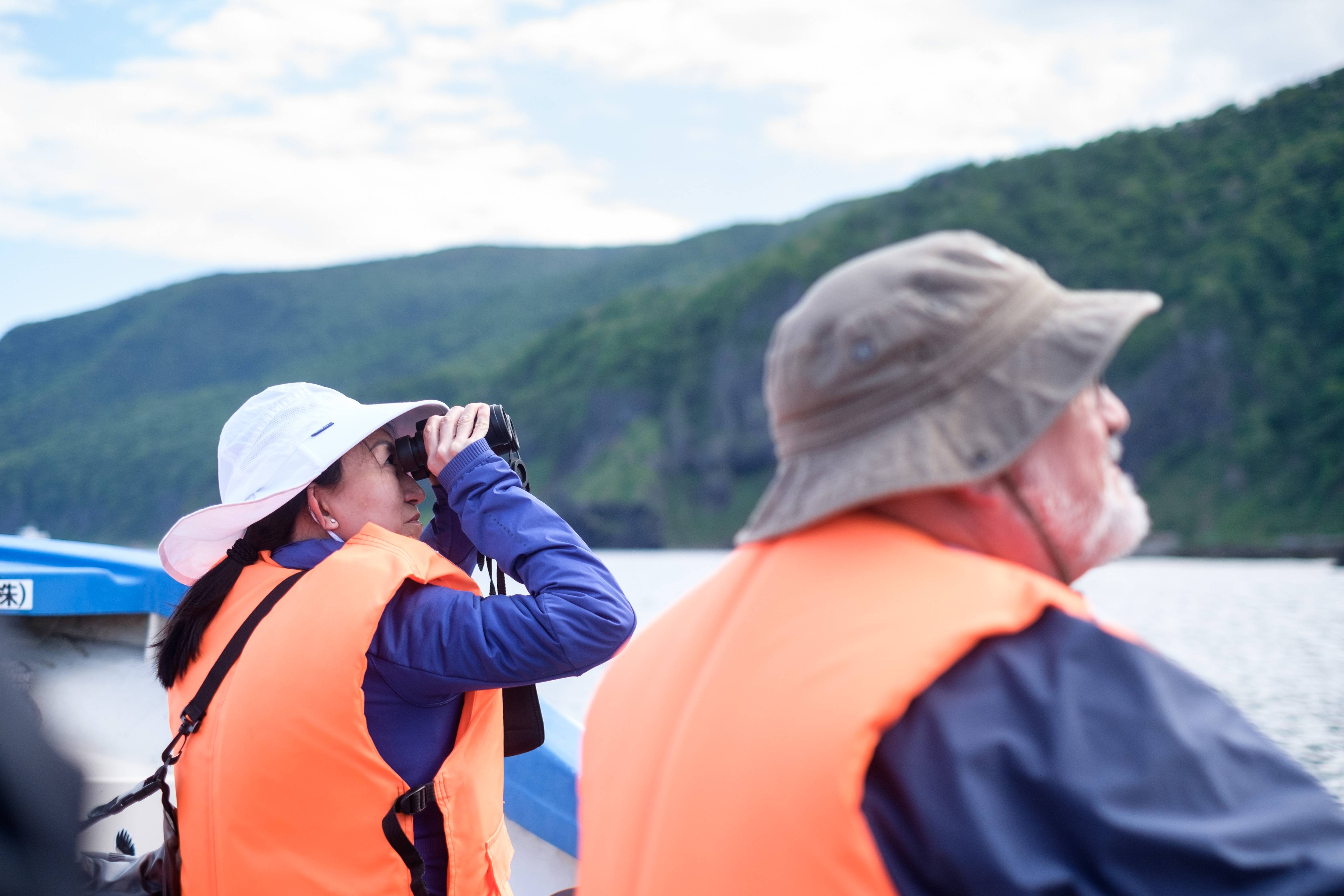 A wildlife watcher wearing a life jacket uses binoculars to look for Brown Bear from a small boat
