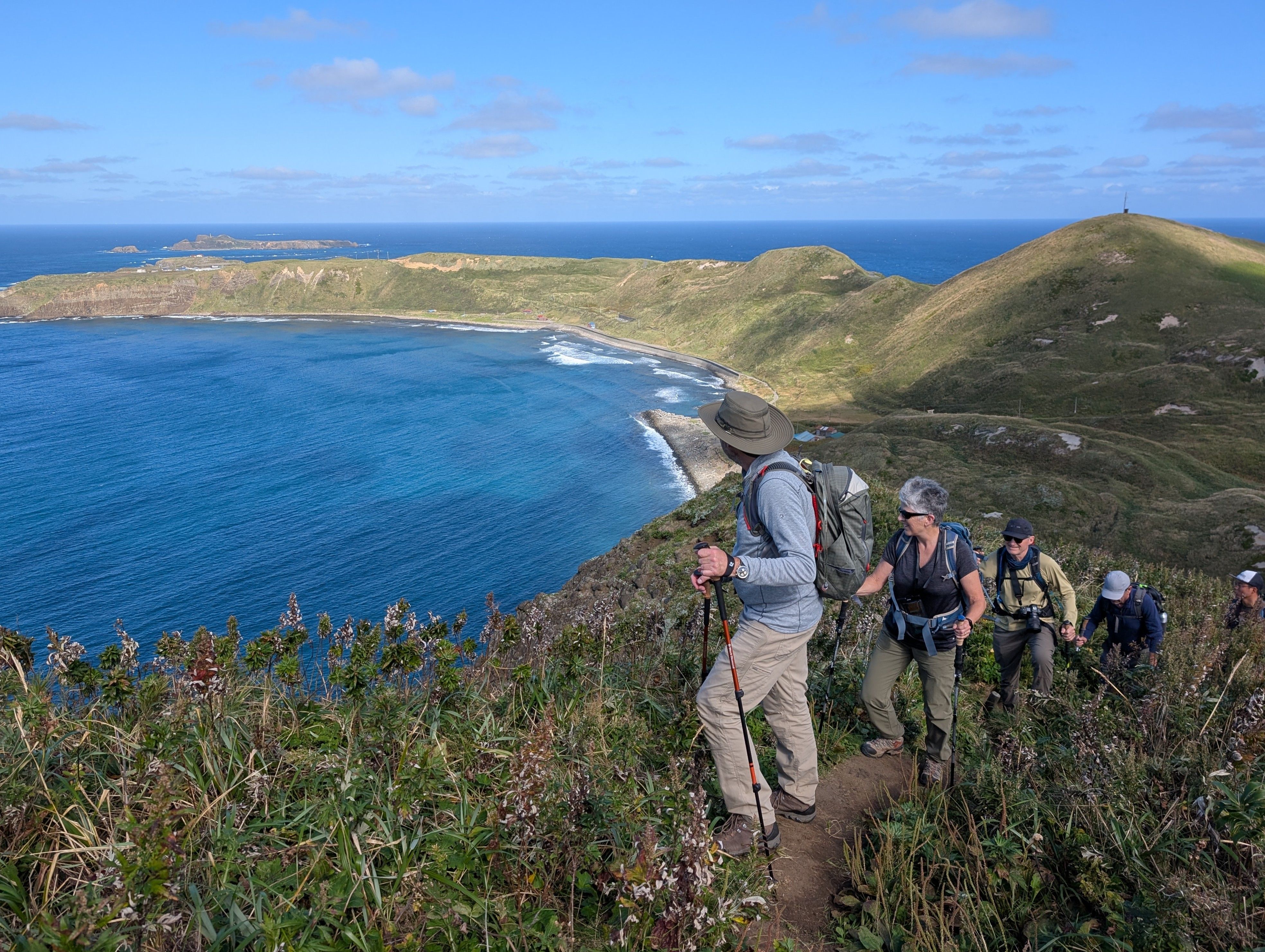 A group of hikers ascend a narrow path on Rebun Island, Hokkaido. They are looking back at a beautiful ocean bay behind them. It is a sunny day and the ocean is reflecting the deep blue colour of the sky.