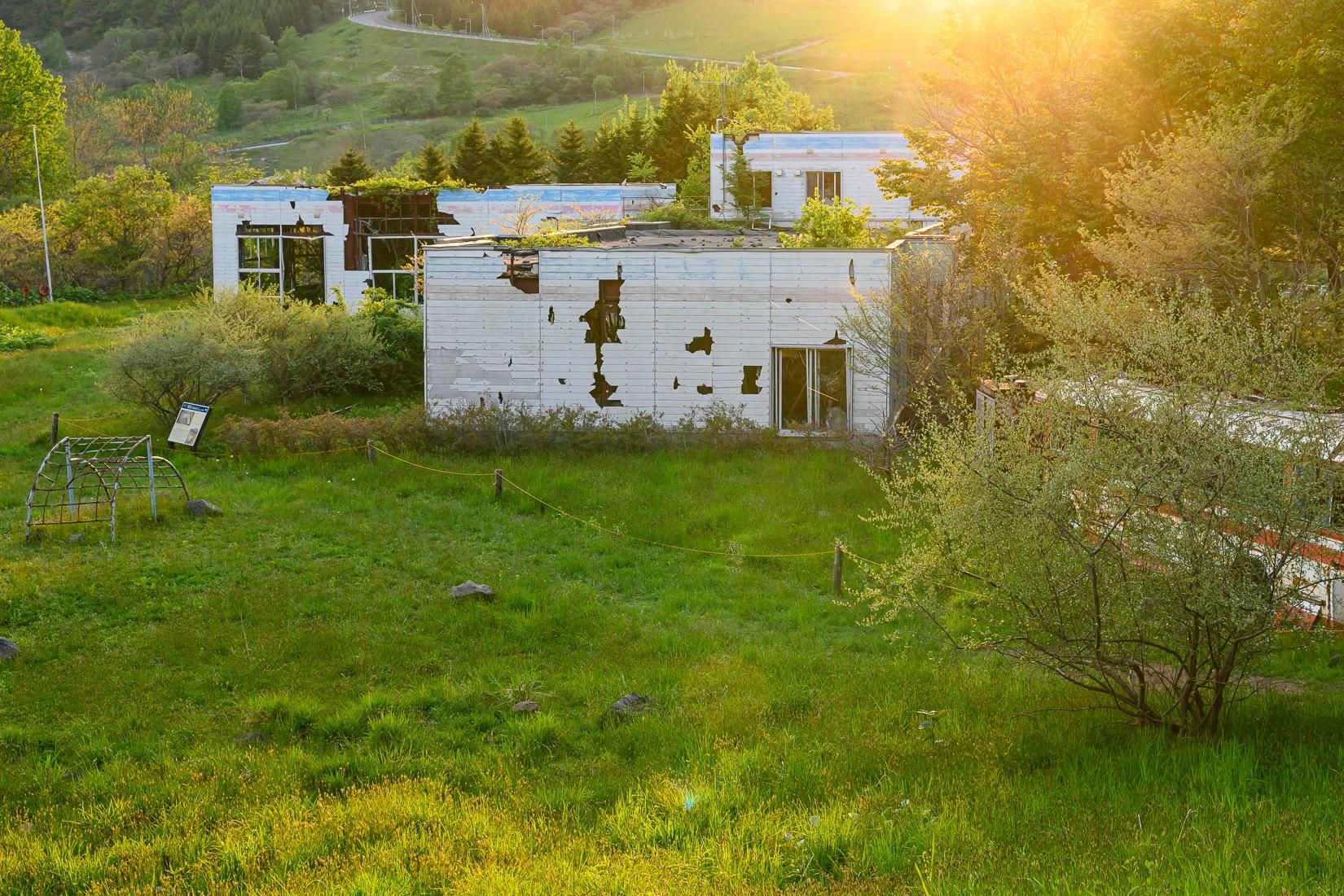 Sunlight illuminates the ruins of a kindergarten damaged by the eruption of Mt. Usu near Lake Toya. The white buildings feature large holes and collapsed sections, now surrounded by lush green grass and trees in a peaceful, reclaimed landscape.