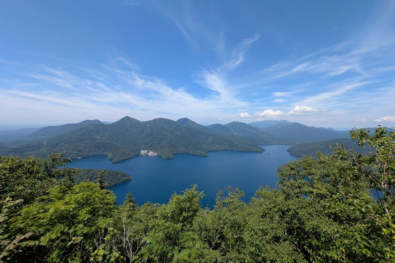 A scenic view from Mt. Hakuunzan overlooking Lake Shikaribetsu, Hokkaido. The image captures the serene blue water surrounded by lush green mountains under a bright sky.