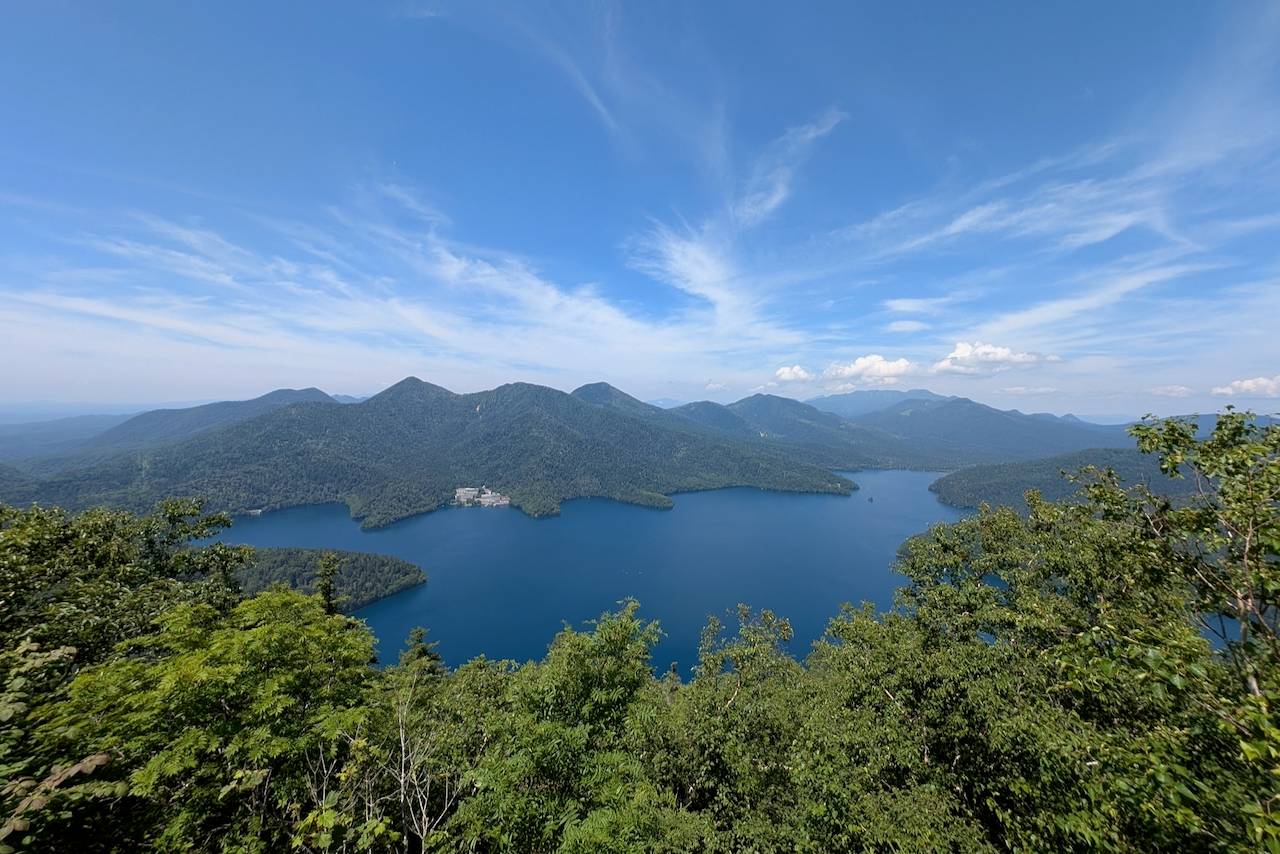 A scenic view from Mt. Hakuunzan overlooking Lake Shikaribetsu, Hokkaido. The image captures the serene blue water surrounded by lush green mountains under a bright sky.