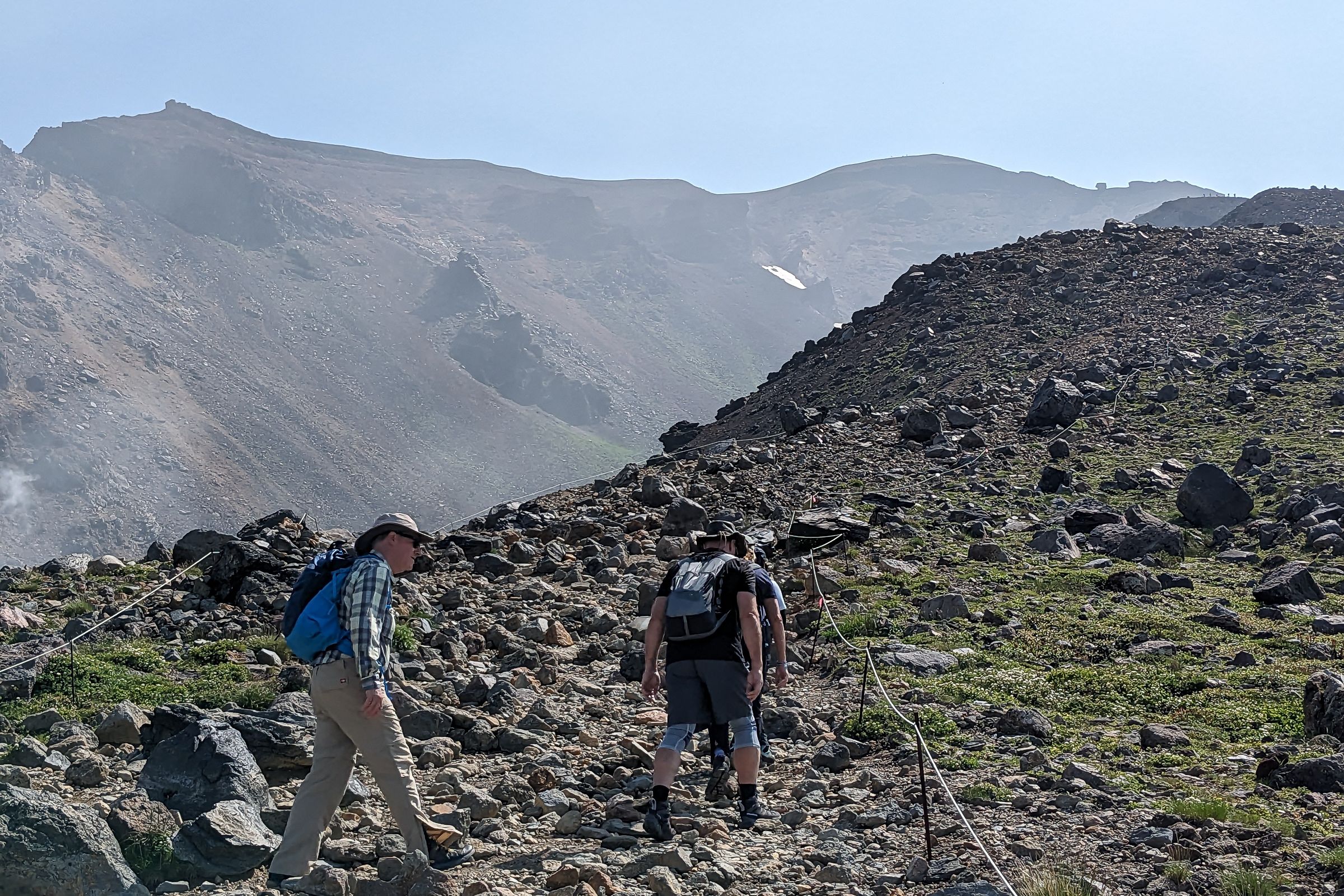 A group of three hikers climb up a rocky path on the way to the summit of Mt. Asahidake. It is a bright sunny day.