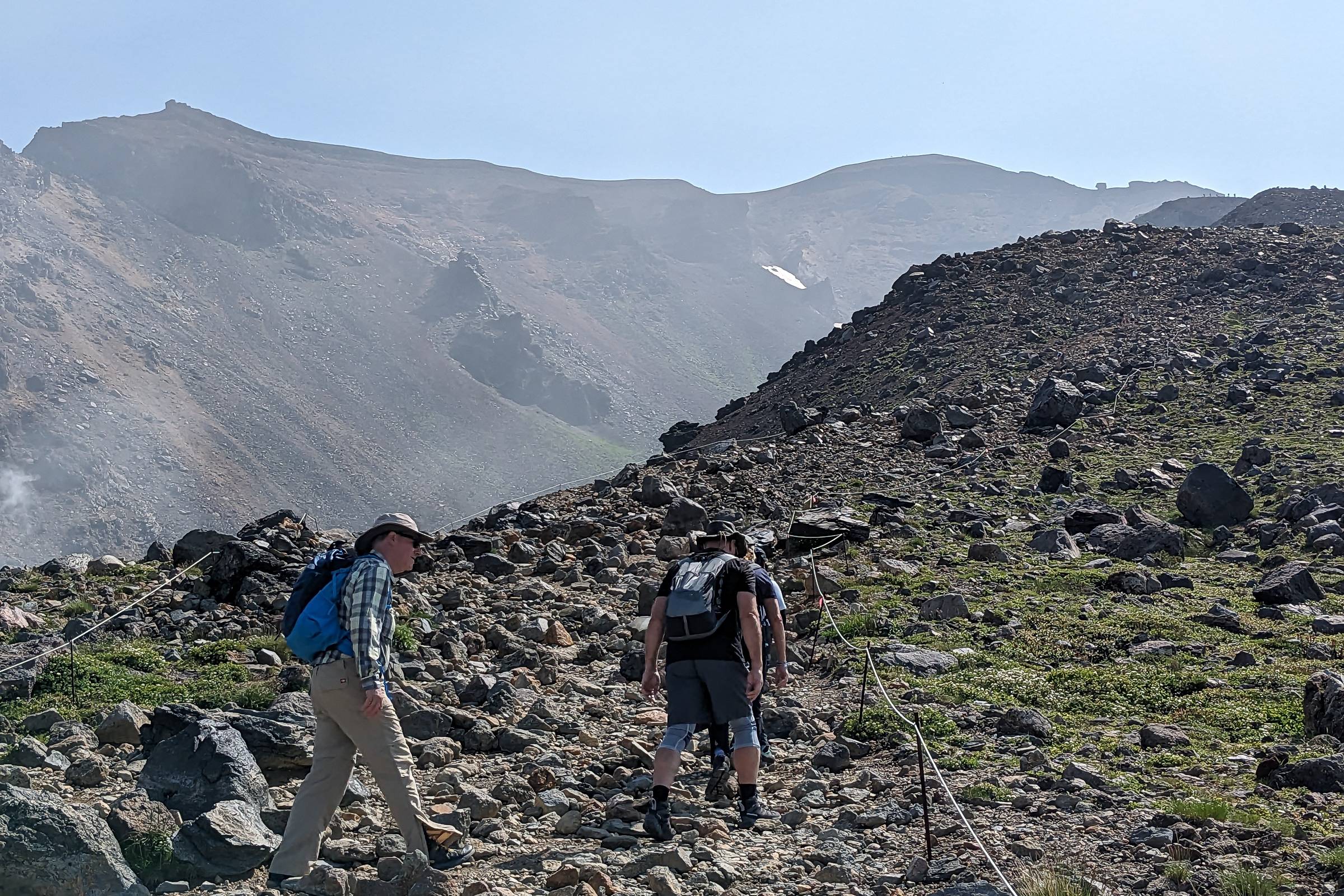 A group of three hikers climb up a rocky path on the way to the summit of Mt. Asahidake. It is a bright sunny day.