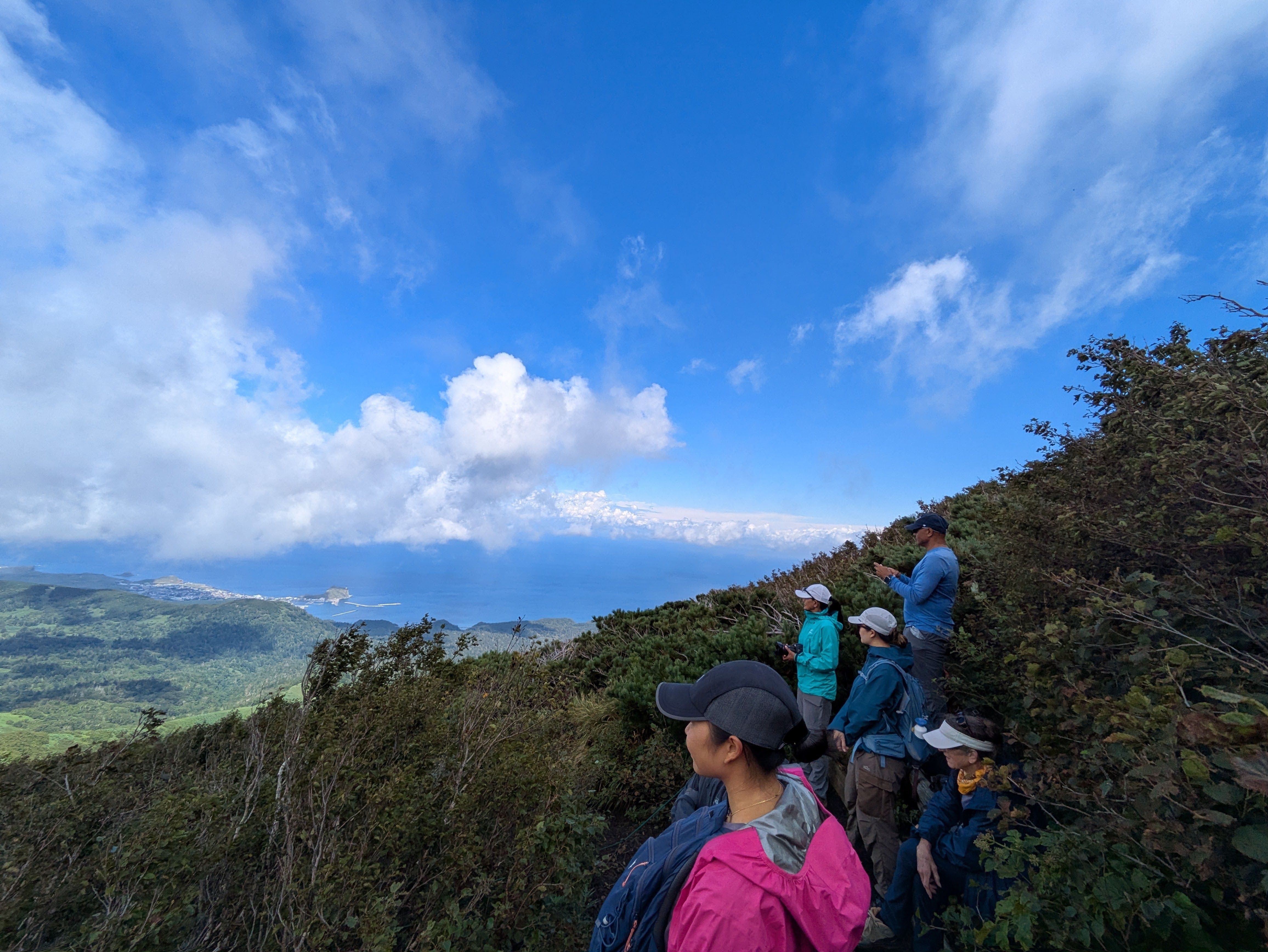 A group of hikers ascend a hiking path on Rishiri-Pon, Mt. Rishiri, Hokkaido. The clouds are clearing from the land below them and the island's forests and the ocean beyond are both visible.