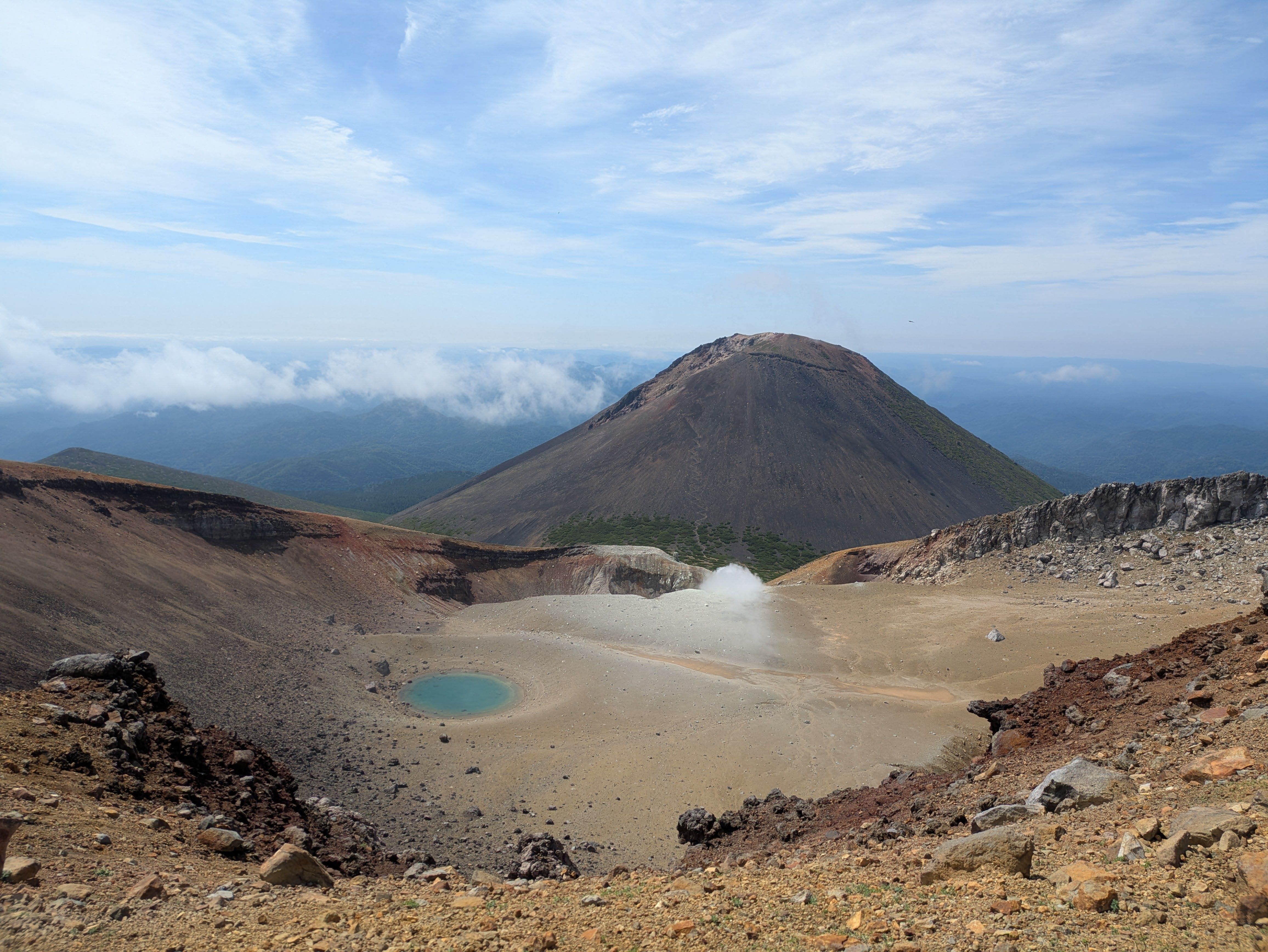 A photograph of the volcanic crater at Mt. Meakan in Hokkaido. A small pond full of bright blue volcanic water is visible in the centre, while a crater with steam rising from it is visible in the midground. The summit of Mt. Akan-Fuji rises in the background.