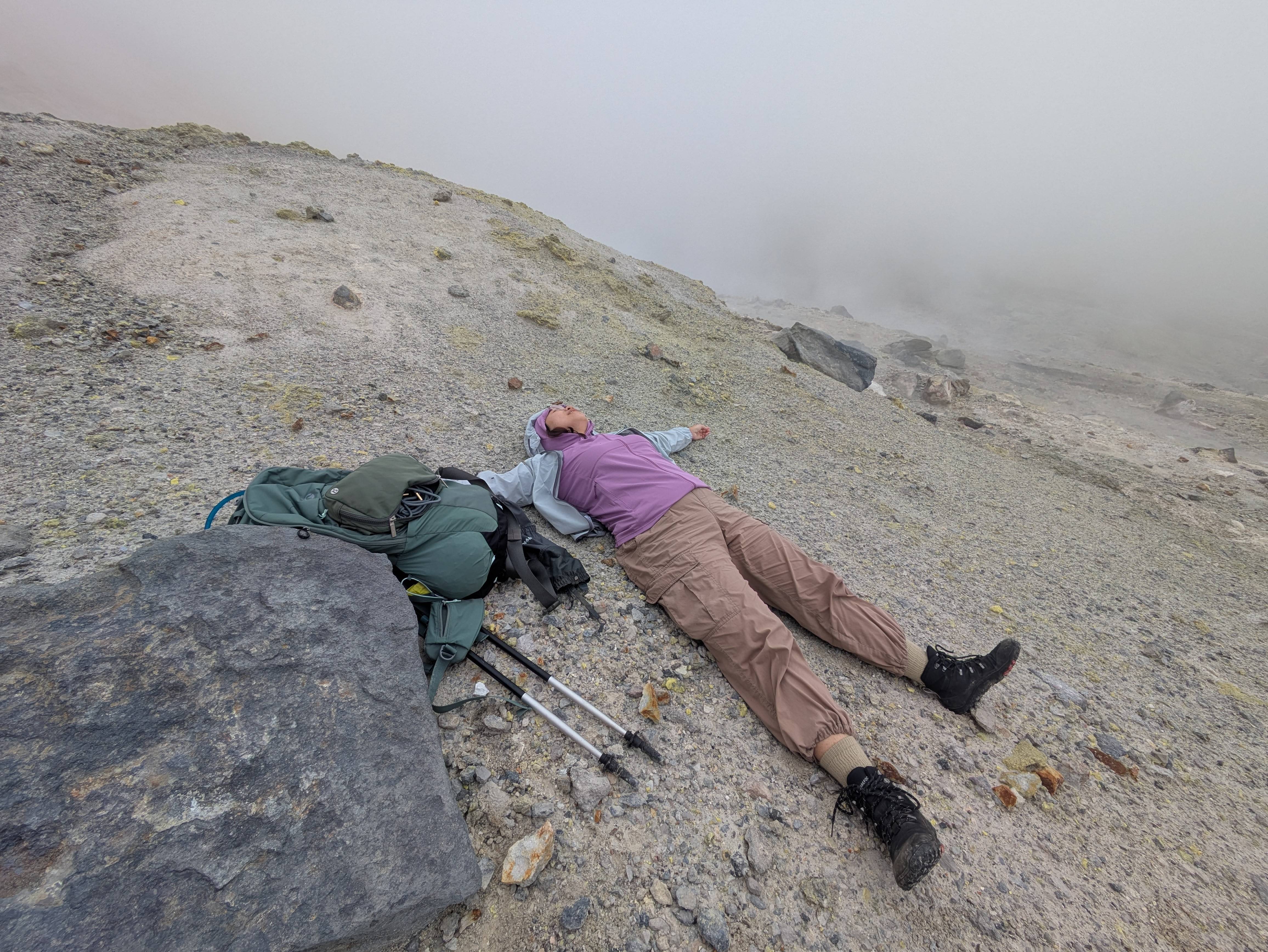 A female hiker lies on the gravel inside Ansei Crater on Mt. Tokachi, Hokkaido, Japan. The scene behind her is cloudy, either with fog or volcanic vents.