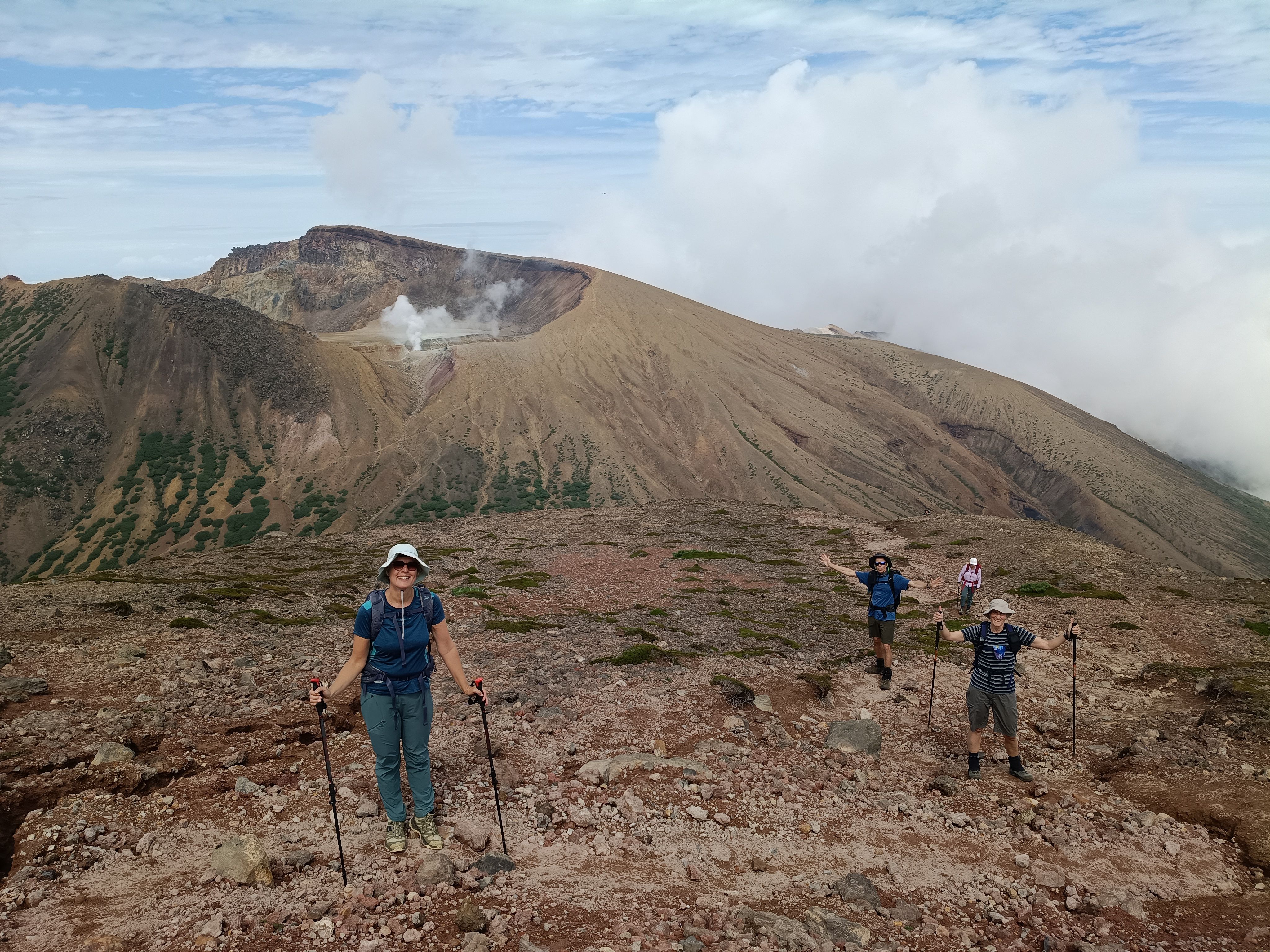 A group of four hikers climb a trail on Mt. Akan-Fuji, Hokkaido. They are all smiling at the camera. Some of the hikers towards the back of the group have their hands raised joyfully. The steaming crater of Mt. Meakan is visible in the background.