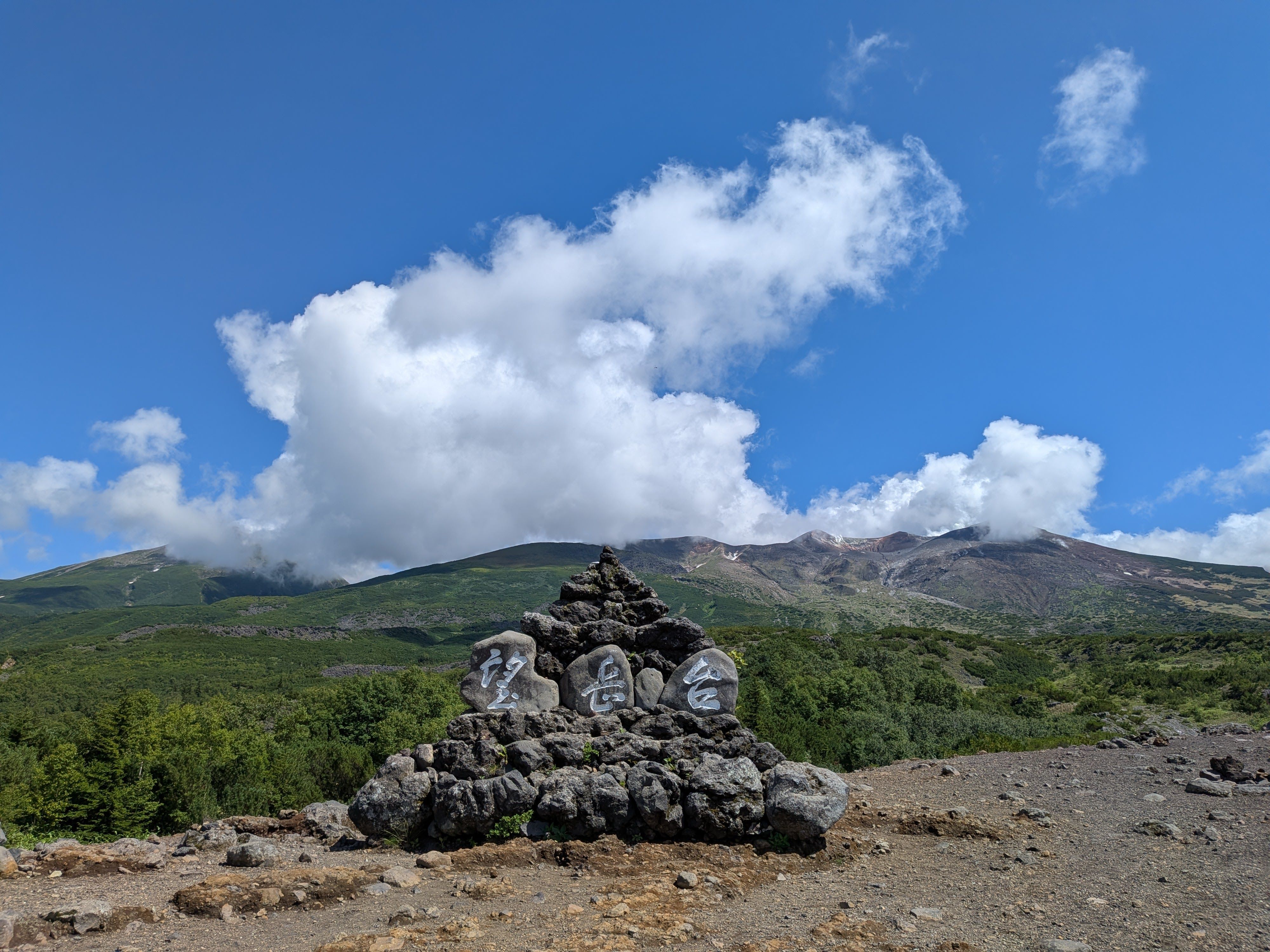 A photograph of Mt. Tokachi in Hokkaido. Steam emerges from the volcano's summit in the distance. A rocky triangle in the foreground is decorated with painted rocks that read "Observatory" in Japanese characters.