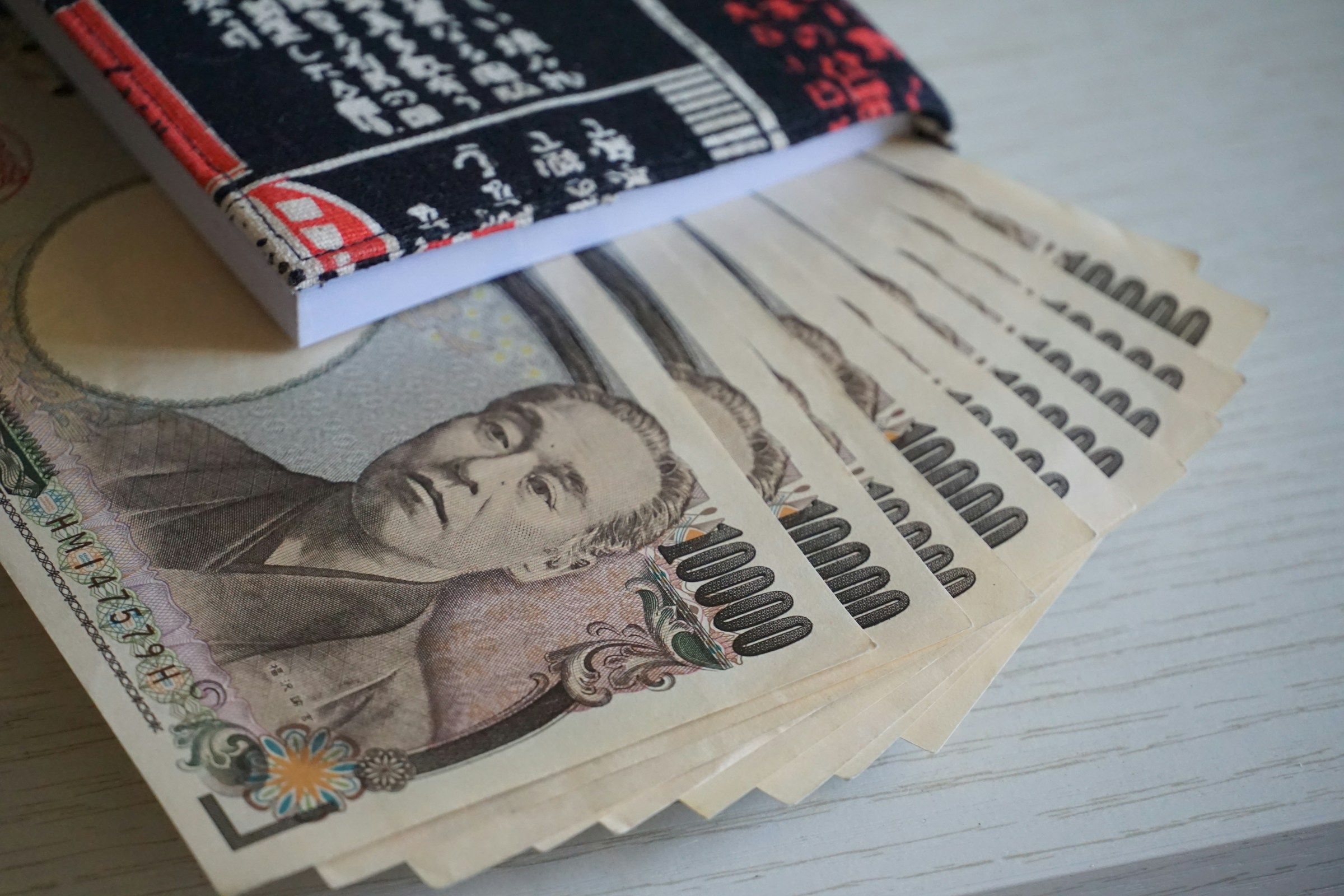 A fanned-out stack of several Japanese 10,000 yen banknotes rests on a table, with a small notebook in a traditional fabric cover lying on top.