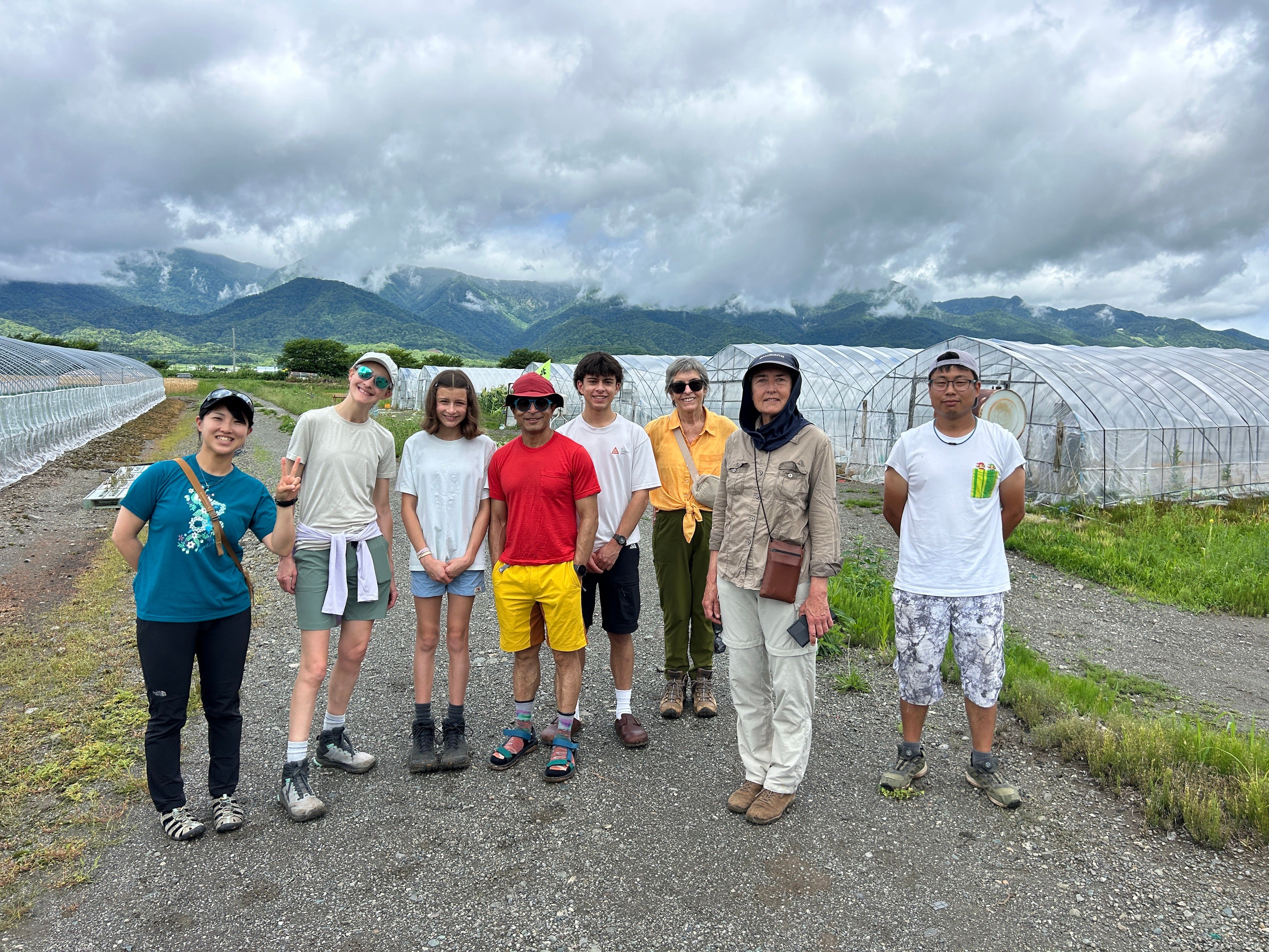 A group of guests stand in a row on a melon farm in Hokkaido, smiling at the camera. Behind them, there are polytunnels growing melons.