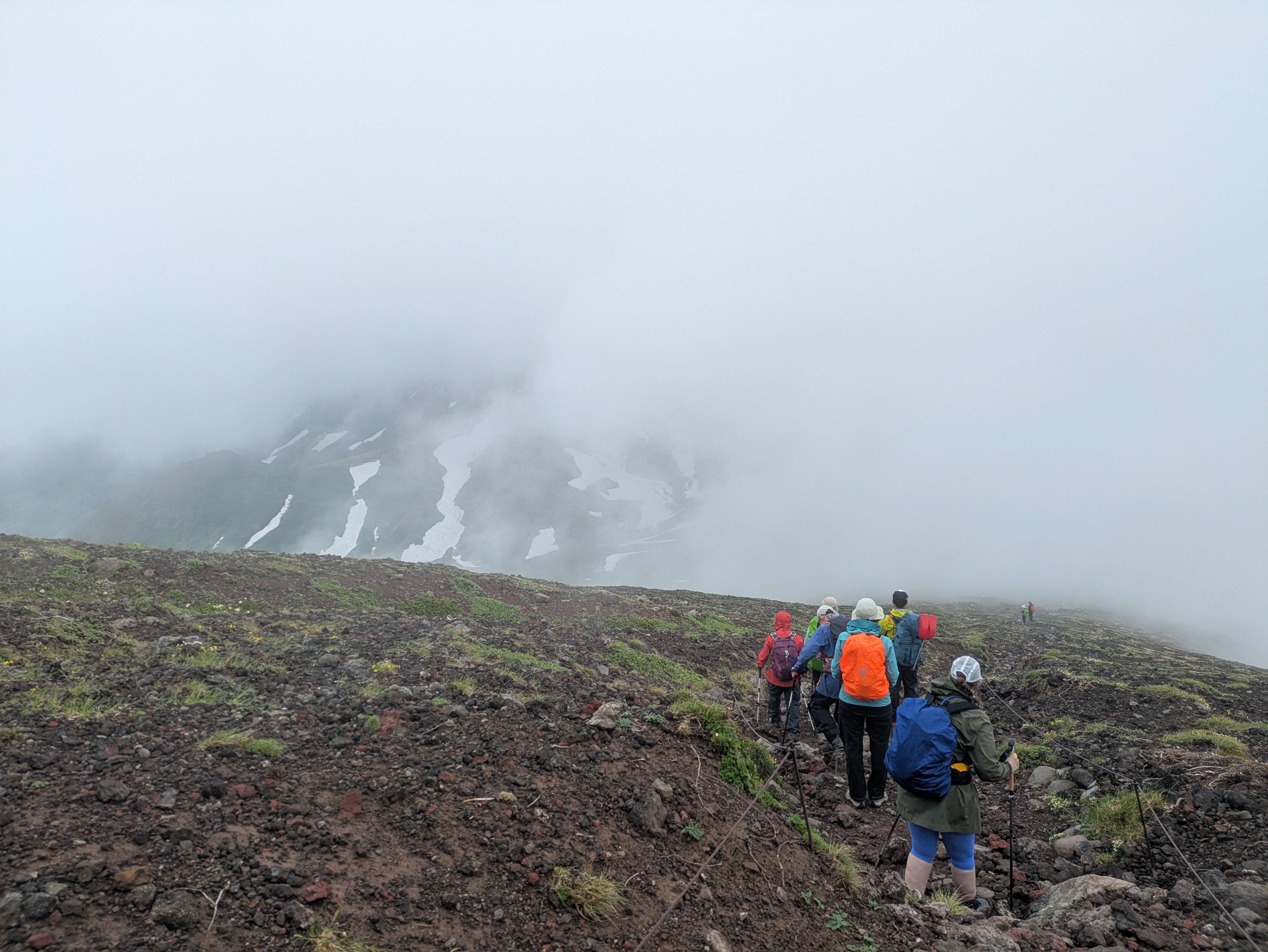 A group of hikers move down a rocky trail on Mt. Asahidake, Hokkaido. It is foggy and the view is partially obscured. Through the clouds, snowfields ate visible on an opposite peak.