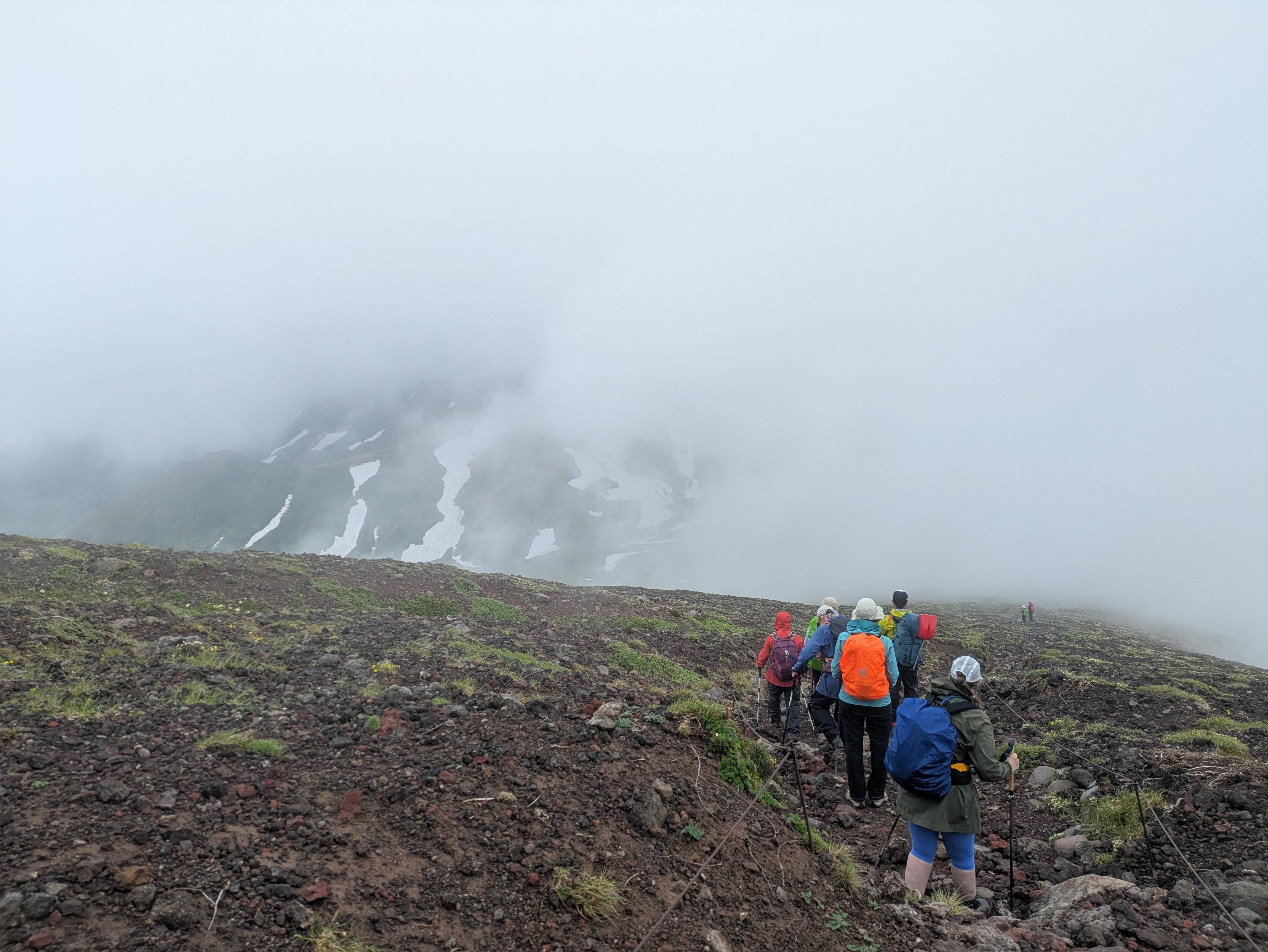 A group of hikers move down a rocky trail on Mt. Asahidake, Hokkaido. It is foggy and the view is partially obscured. Through the clouds, snowfields ate visible on an opposite peak.