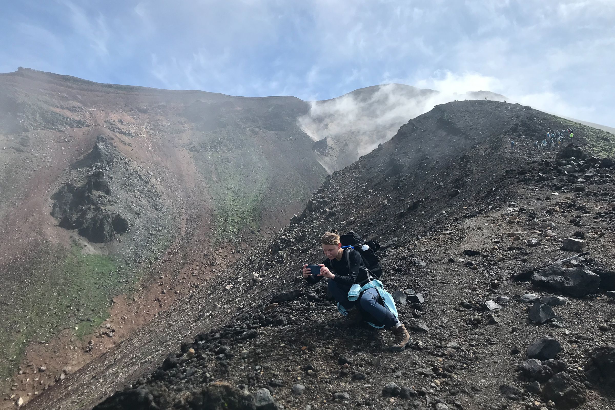 A hiker leans on a mountain slope to take a photograph of the scenery.