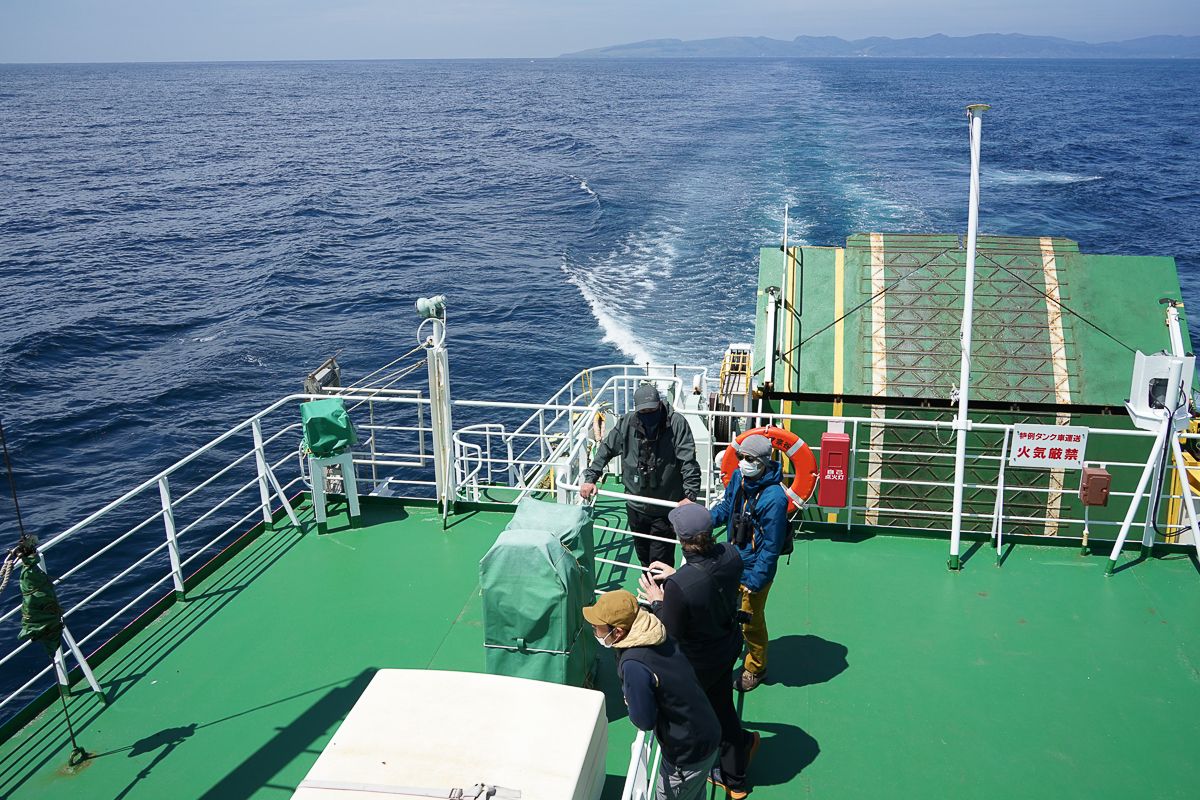 A group of guides gathers on the green deck of the ferry to Rebun Island. The deep blue ocean stretches out, with the boat's wake leading toward the distant land on the horizon.
