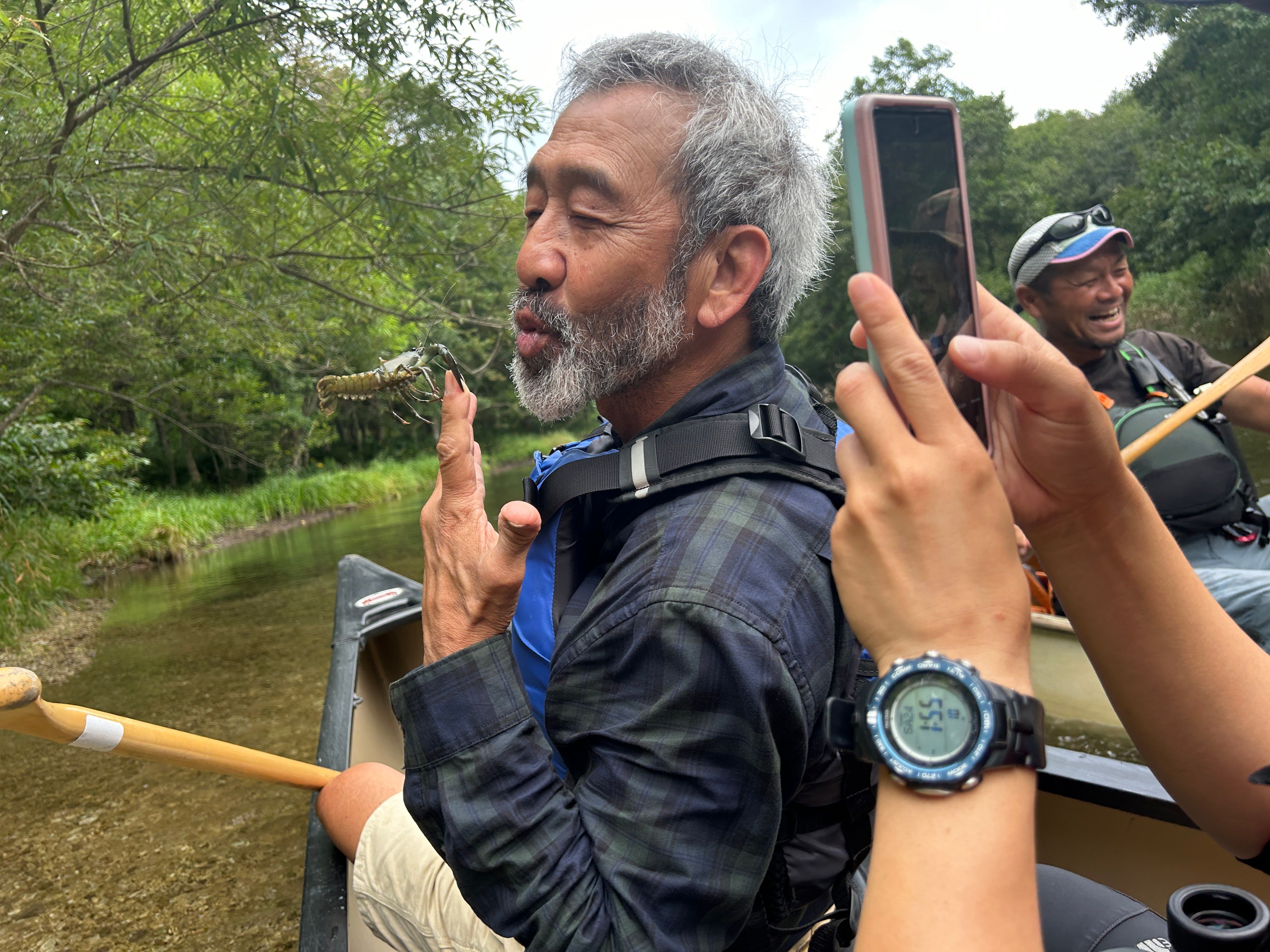 A man jokes around with a crayfish pinching his finger! He is holding it up to his mouth as if to kiss it. Someone in the foreground has raised their smartphone and is taking a photo of the scene.