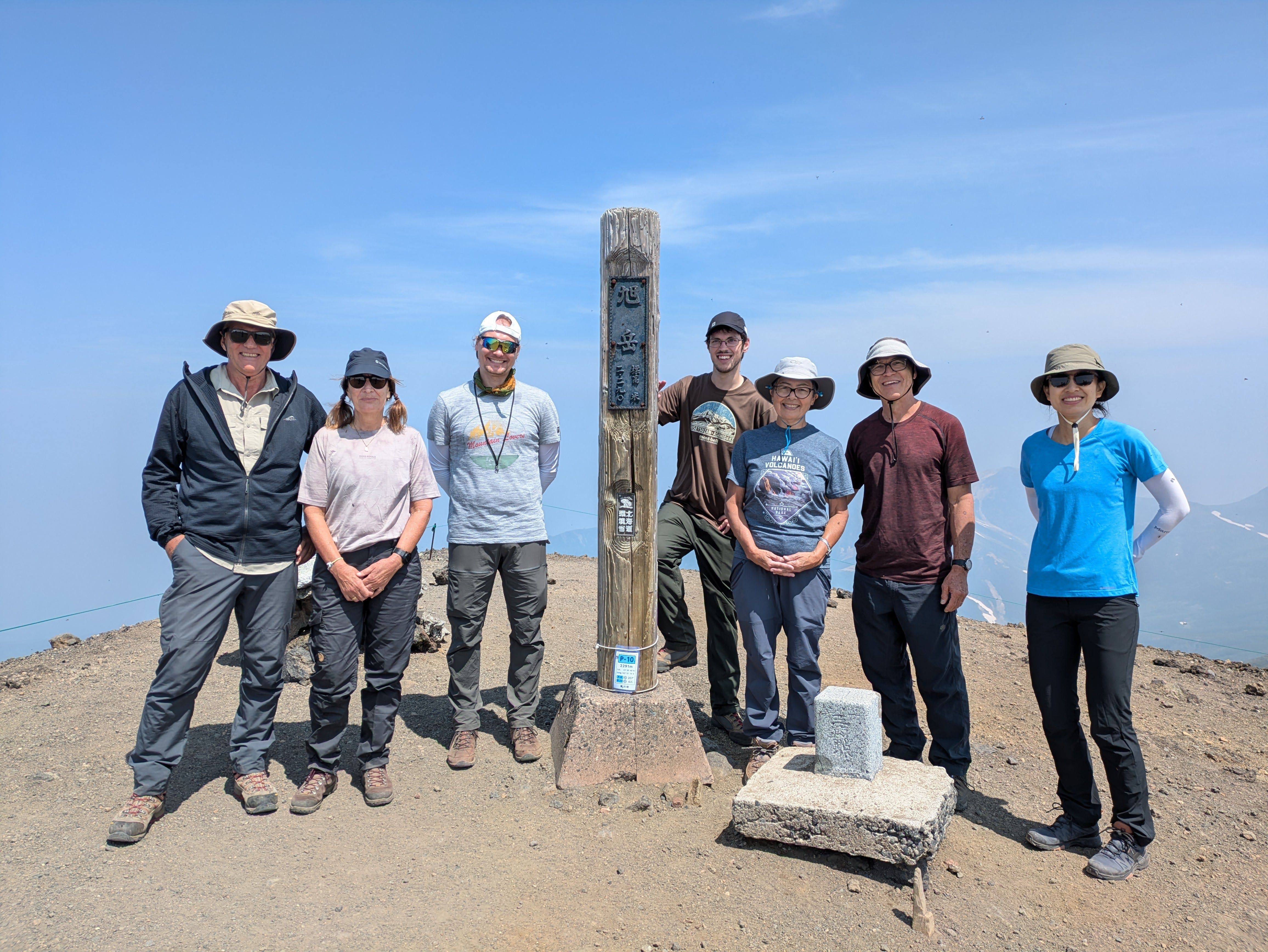 A group of hikers at the summit of Mt. Asahidake in Hokkaido. They are stood around a pole which reads in Japanese "Mt. Asahidake - 2290m"