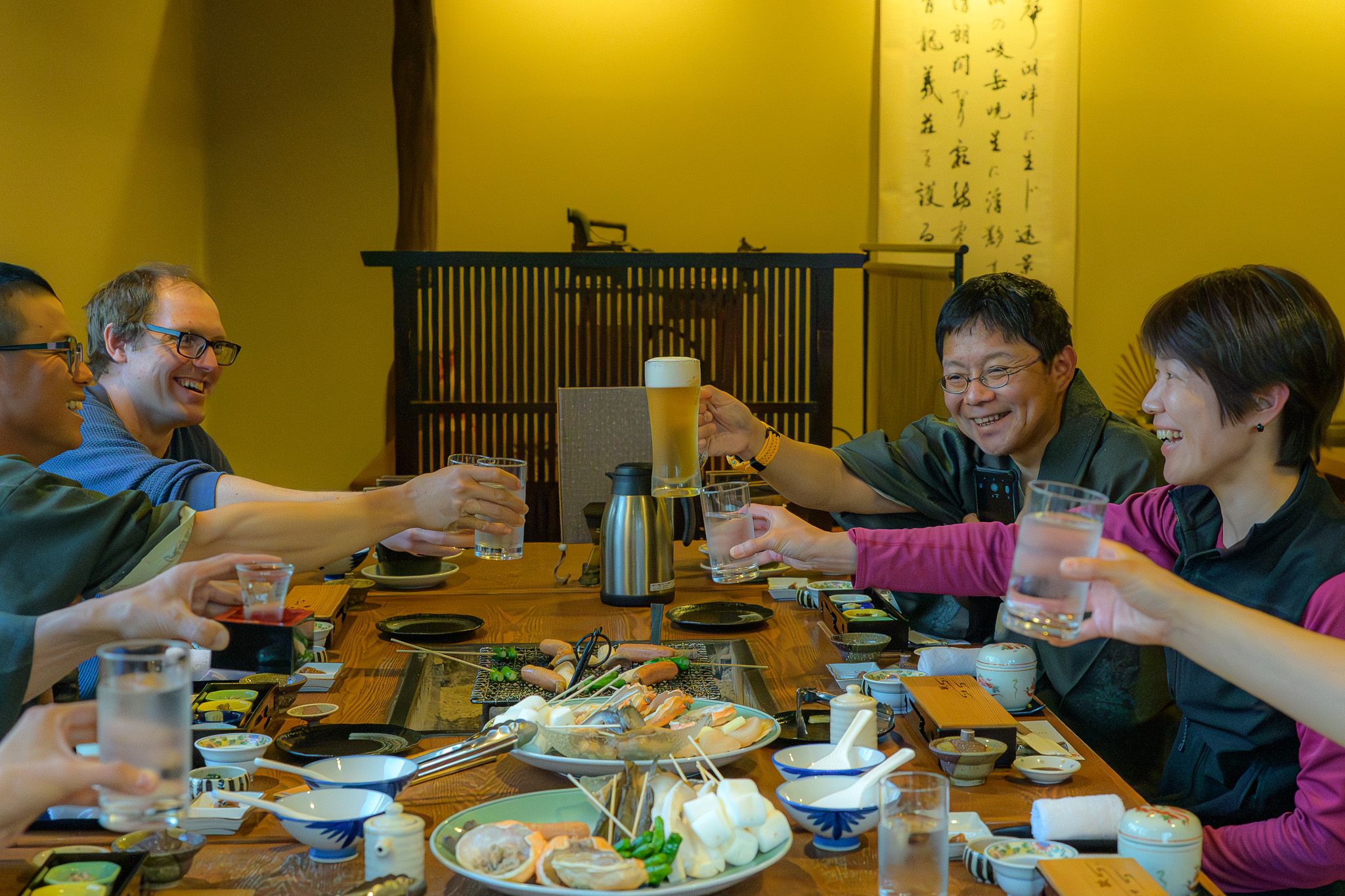 The diverse Adventure Hokkaido staff team shares a meal, happily raising their glasses in a toast around a large wooden table laden with Japanese dishes.