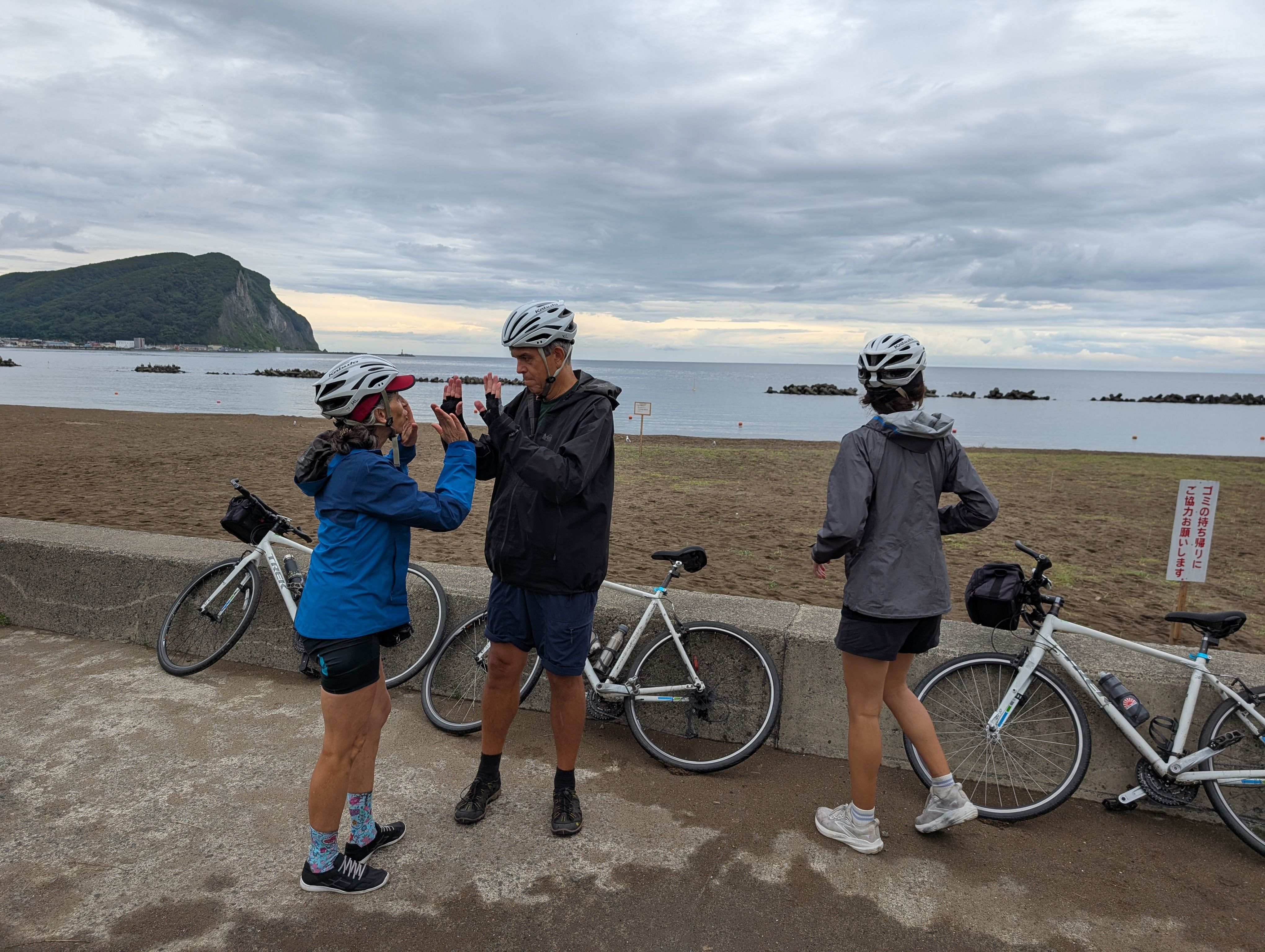 Three cyclists are dismounted near a beach looking out to the Sea of Japan. Two of the cyclists are double high-fiving in celebration.