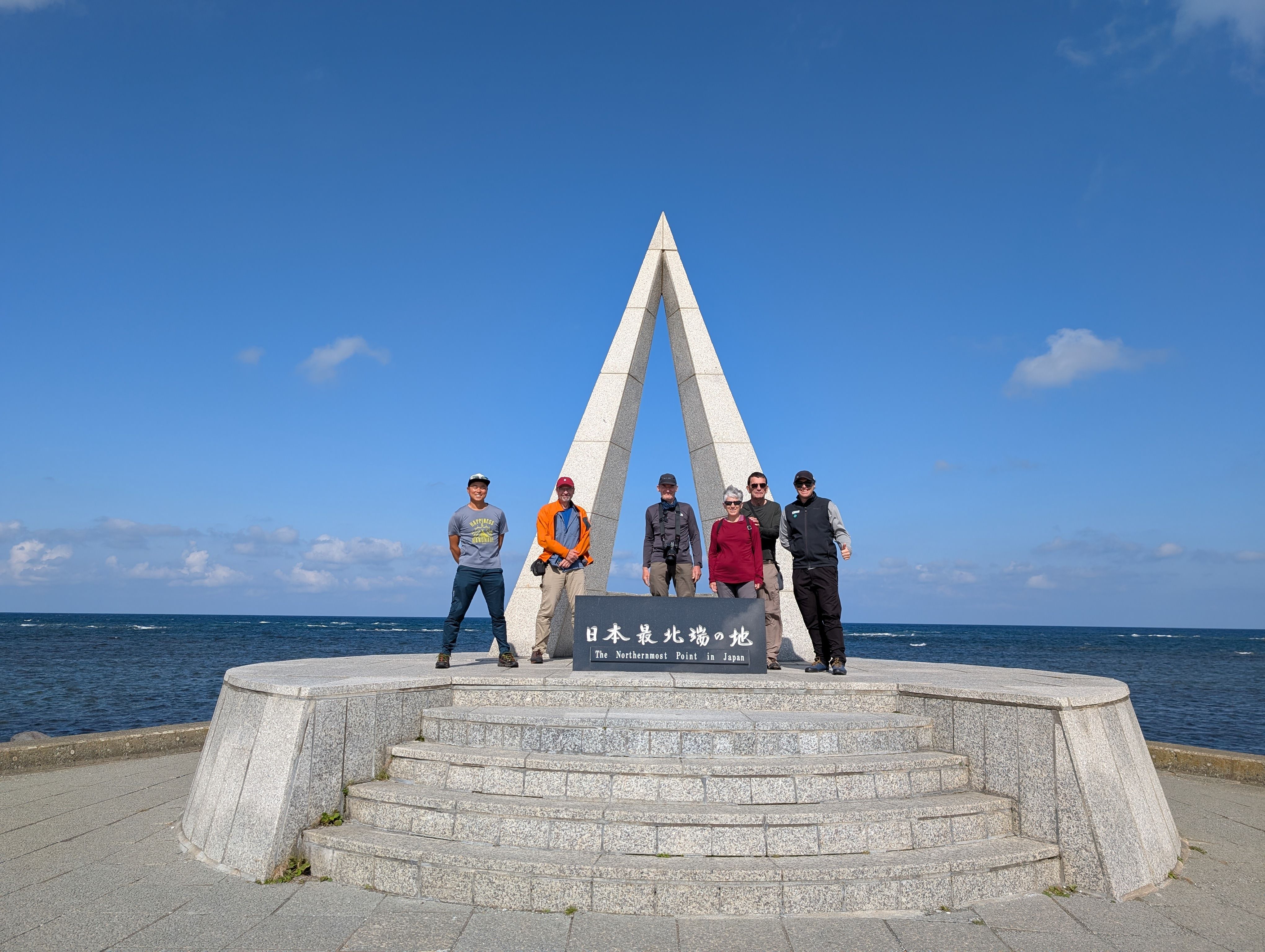 A group of people stand next to a triangular monument in front of the ocean on a sunny day. A sign in front of them reads, "The northernmost point in Japan" in both Japanese and English.