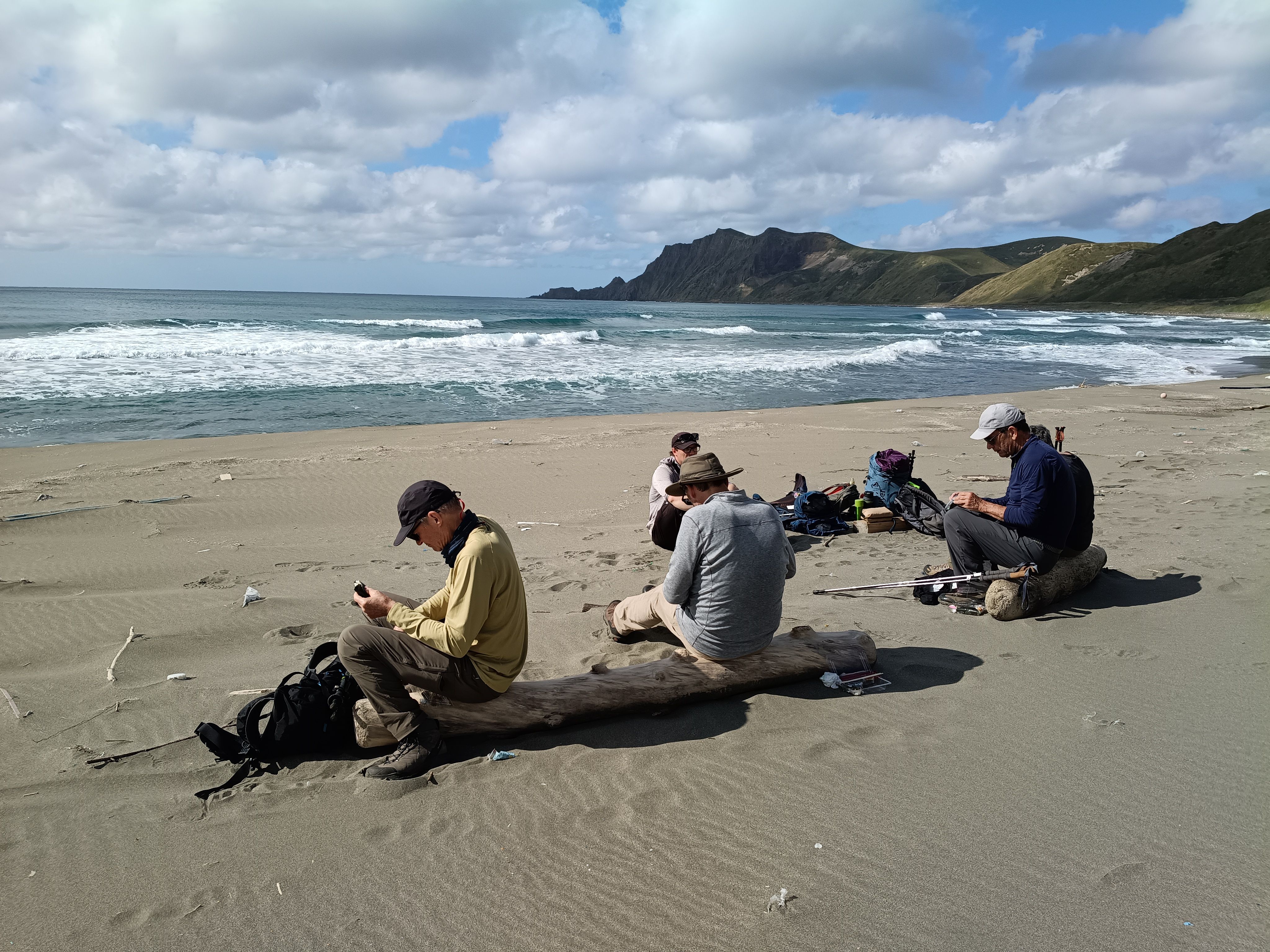 A group of hikers take a break on the beach of Rebun Island, Hokkaido. They sit on driftwood in the sand. Some are looking at smartphones, others eating rice balls. It is a clear day, but it also appears to be quite windy.