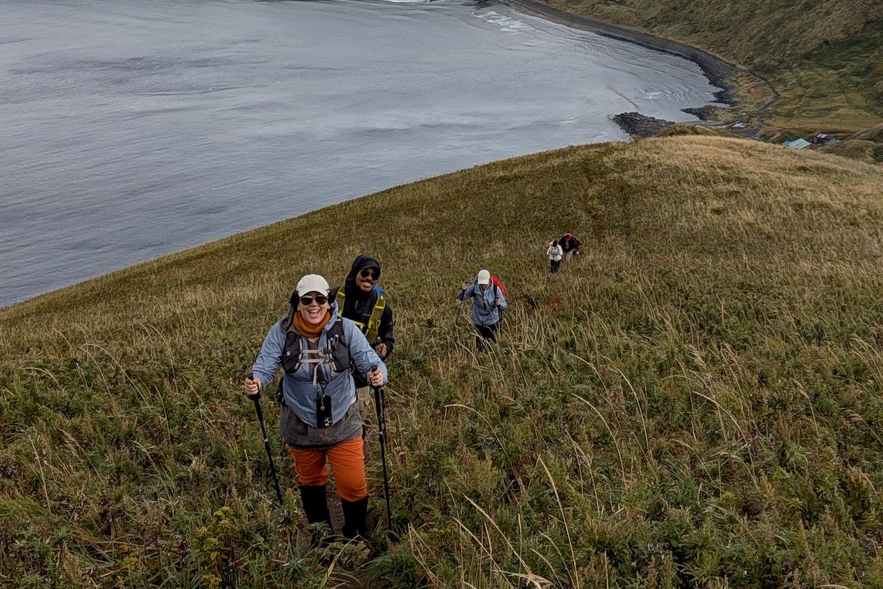 A group of smiling hikers ascending a steep, grassy hillside on the Misaki Meguri trail on Rebun Island, Hokkaido. In the foreground, a woman beams joyfully while using trekking poles. Behind the hikers, the calm ocean and a curving shoreline stretch out below.