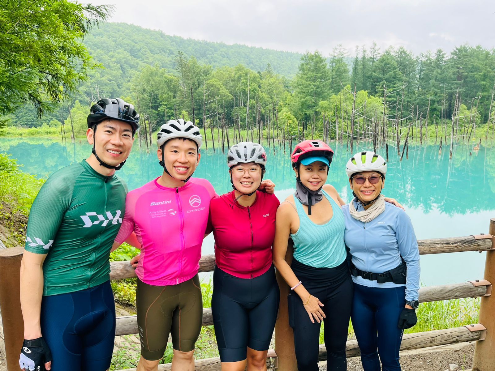 Five cyclists in their helmets and cycling gear have their arms around each other and smile at the camera in front of the Blue Pond in Hokkaido.