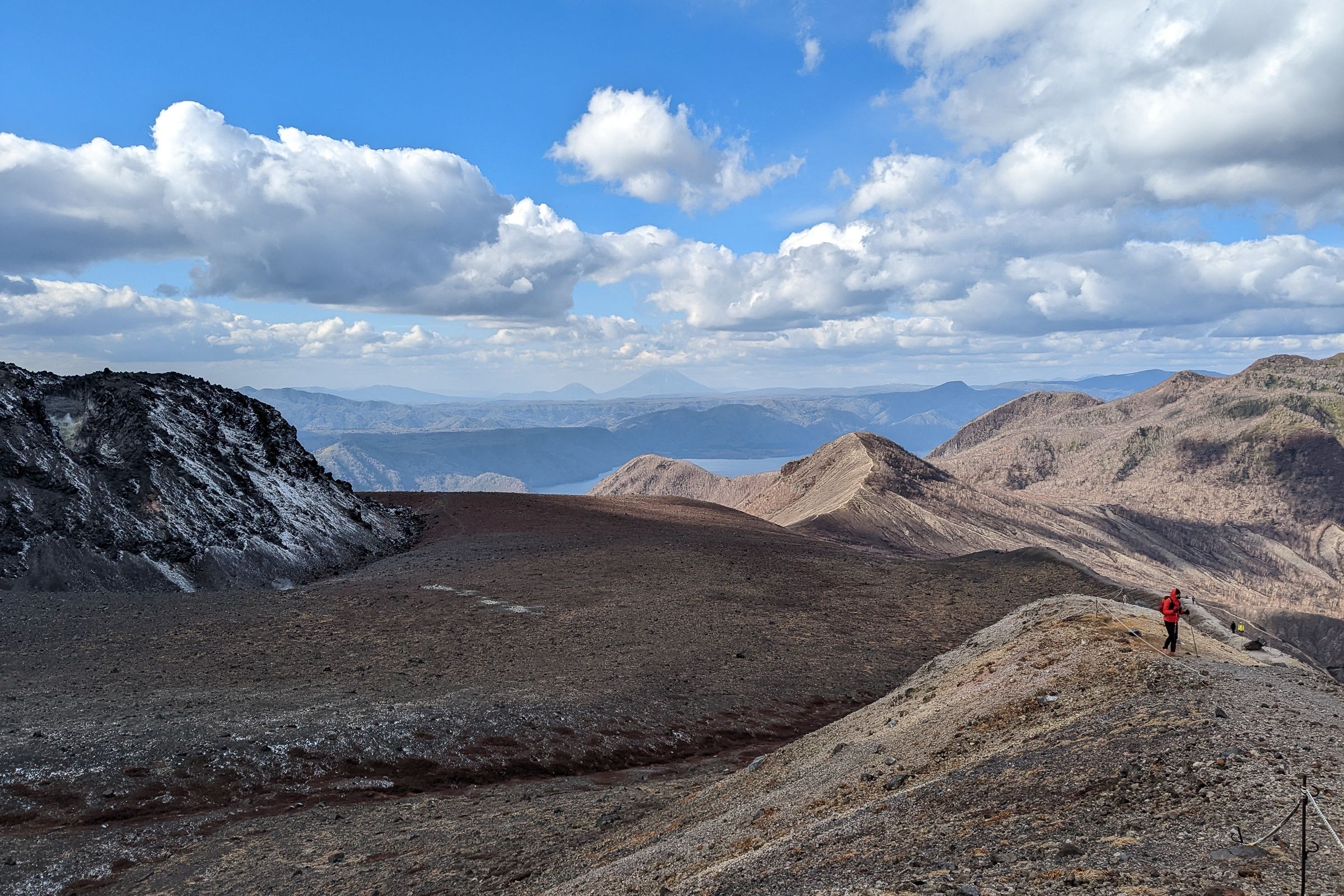 A hiker wearing a red rain jacket traverses Mount Tarumae's outer rim trail, with dramatic views of the volcanic crater and Lake Shikotsu in the background.