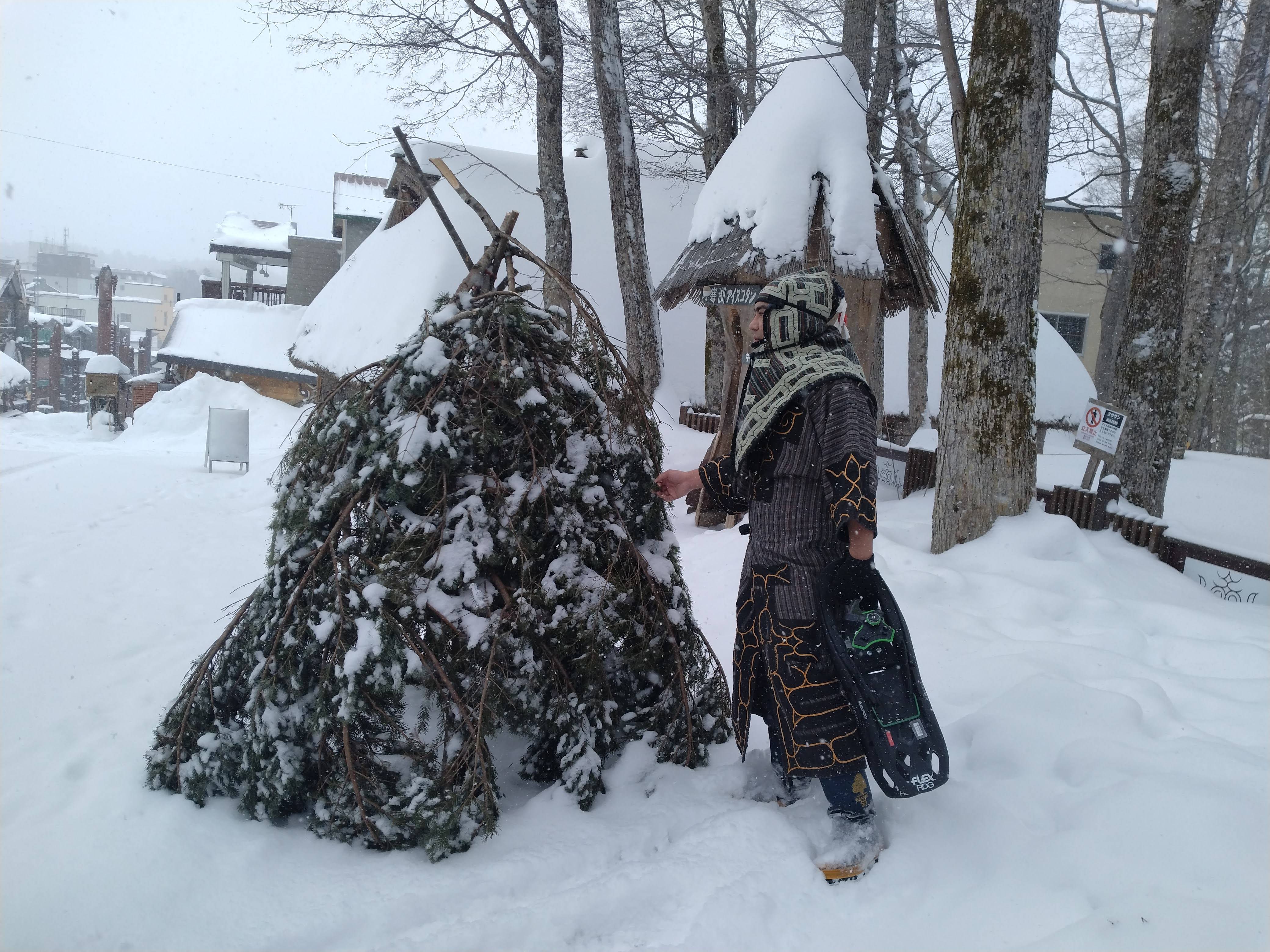 An Ainu man in traditional Ainu clothing stands next to a snow-covered shelter made of fir tree branches. It is snowing heavily.