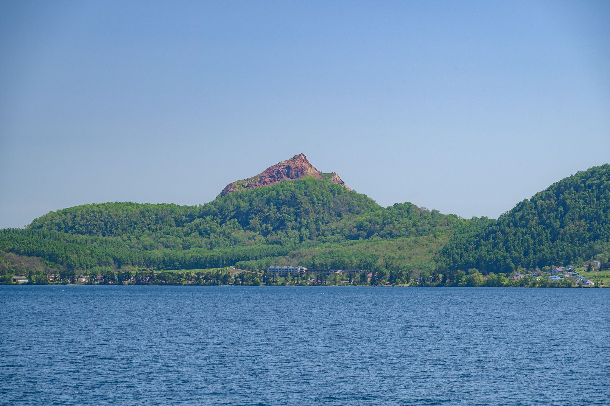 A vibrant, wide-angle shot of Lake Toya, with Mt. Showashinzan dominating the background. The young mountain's upper peak is a striking reddish-brown, devoid of foliage, contrasting sharply with the verdant green trees covering its lower slopes and the surrounding hills. In the mid-ground, a scattering of buildings can be seen along the lake's edge, nestled amongst the trees. The foreground is filled with the deep blue waters of Lake Toya under a clear, bright blue sky.