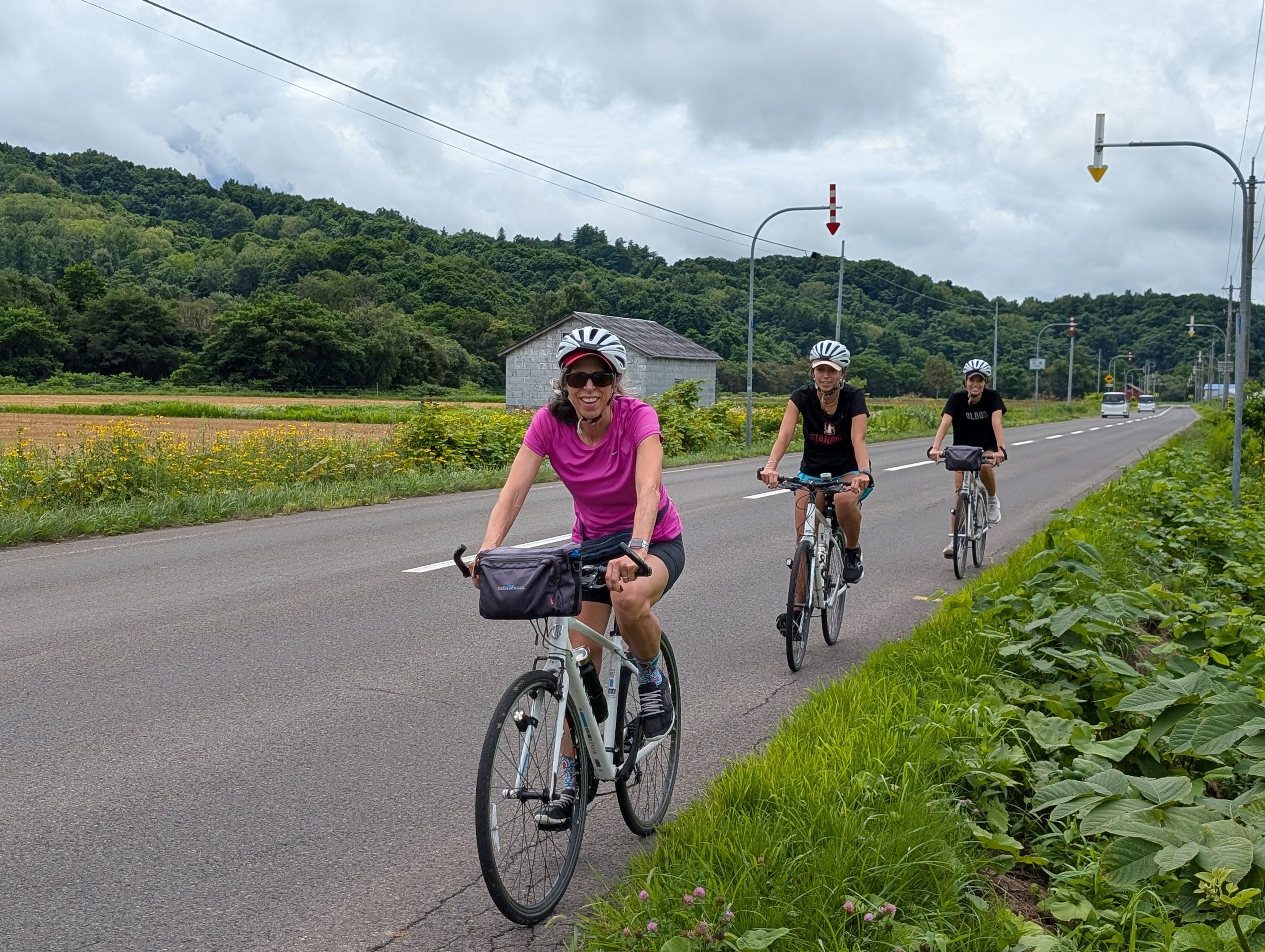 Three cyclists ride along the road in Hokkaido farmland. The woman at the front of the line of cyclists smiles at the camera.