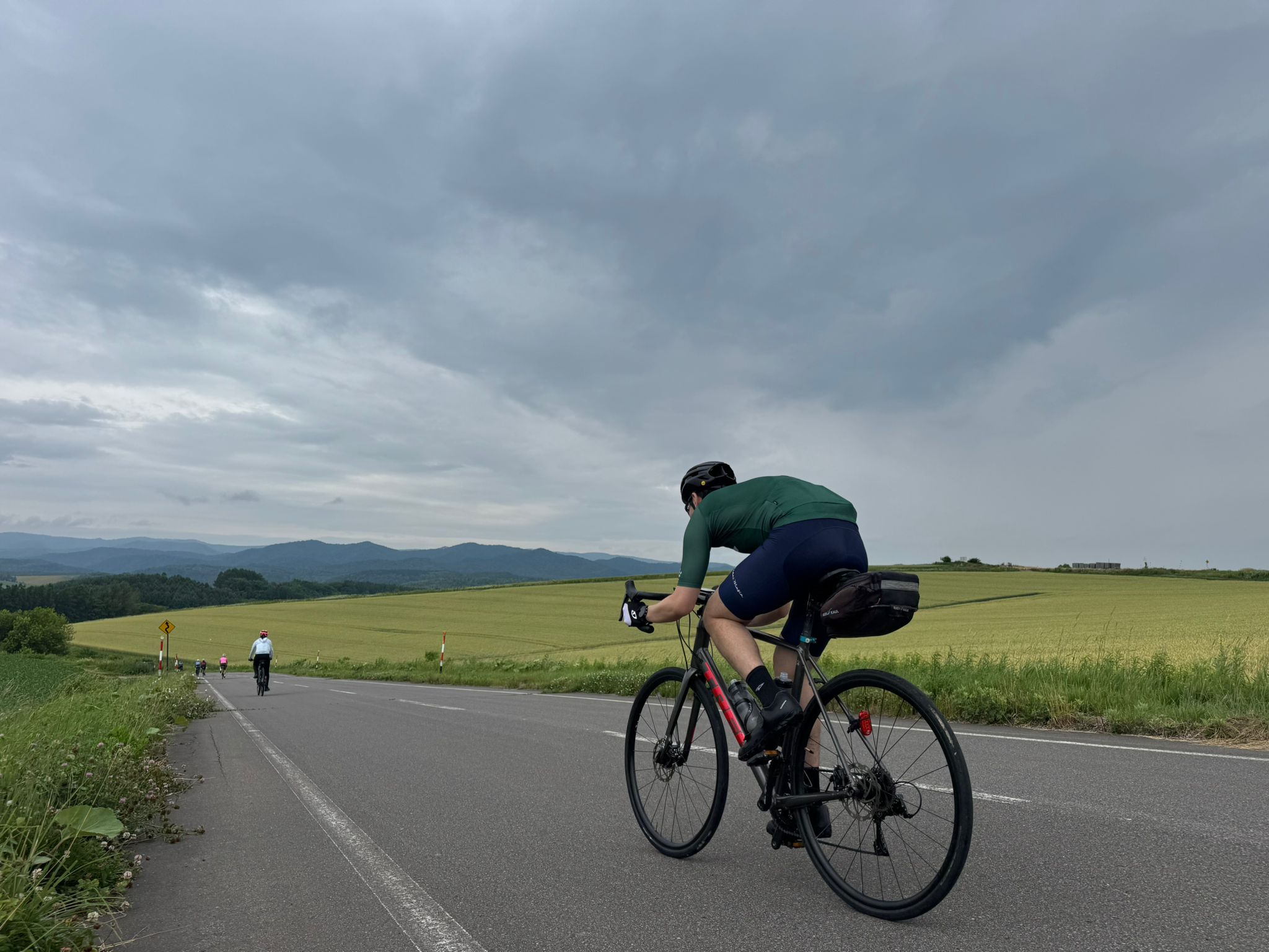 Two cyclists ride downhill in Biei, Hokkaido. There is farmland and vast wheat fields all around them.