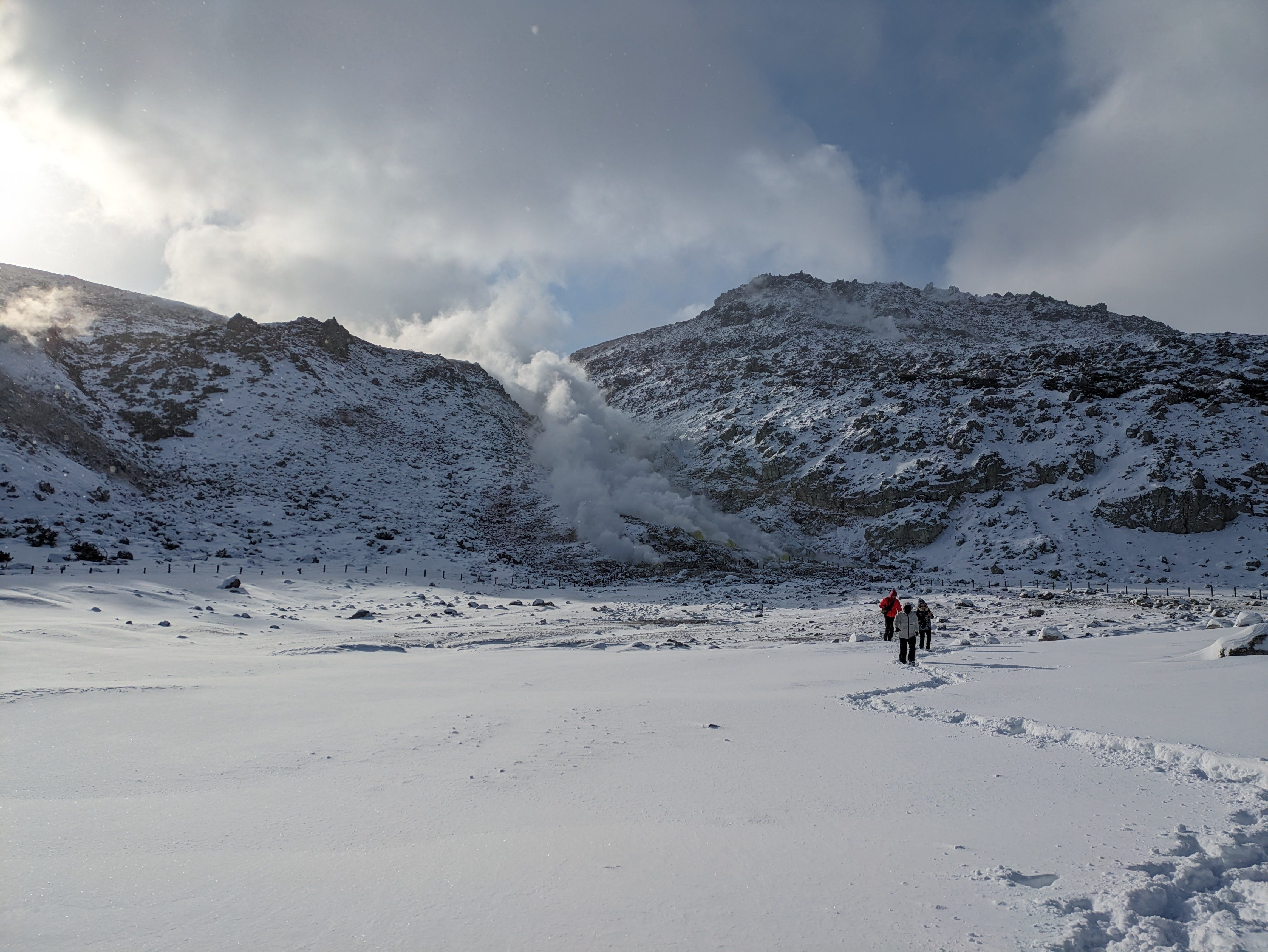 Guests leave a trail of footprints in the snow as they approach Mt. Io, its steam rushing upward towards the clouds in heaven.
