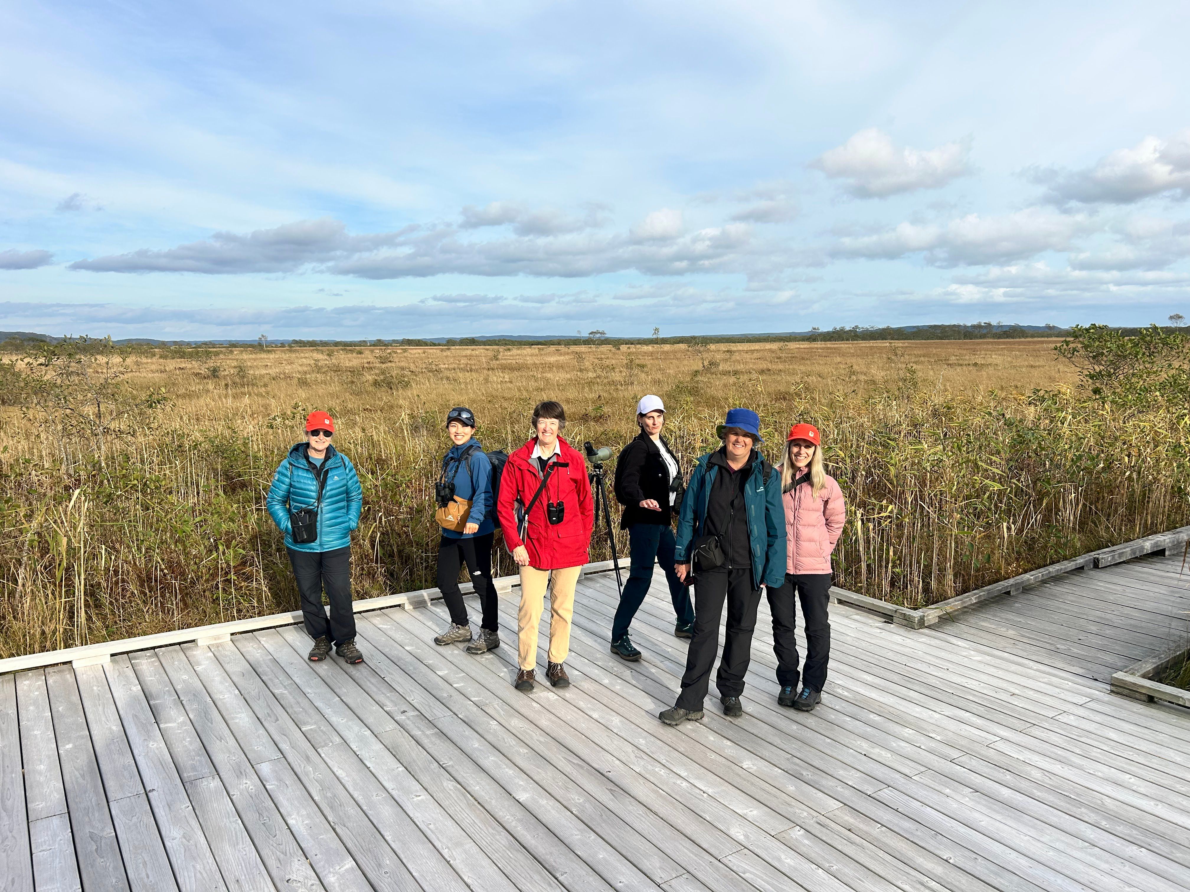A group of hikers on a boardwalk at Onnenai Grassland in Kushiro-Shitsugen National Park, Hokkaido. The grassland is visible behind them in its early autumn colours. It is a sunny day with high, wispy clouds overhead.