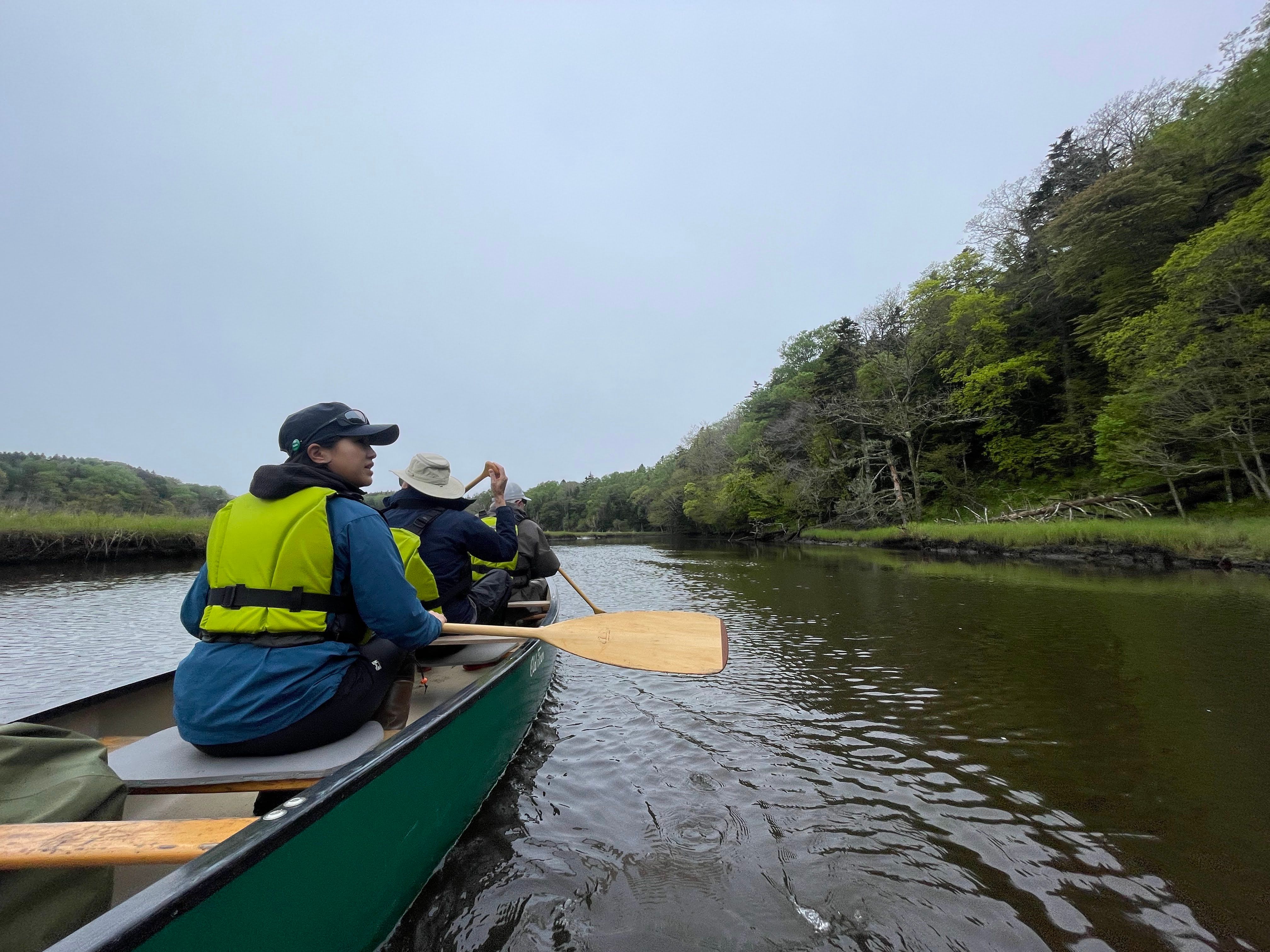 Three people paddle a canoe down the Bettoga river in east Hokkaido. The sky is overcast.