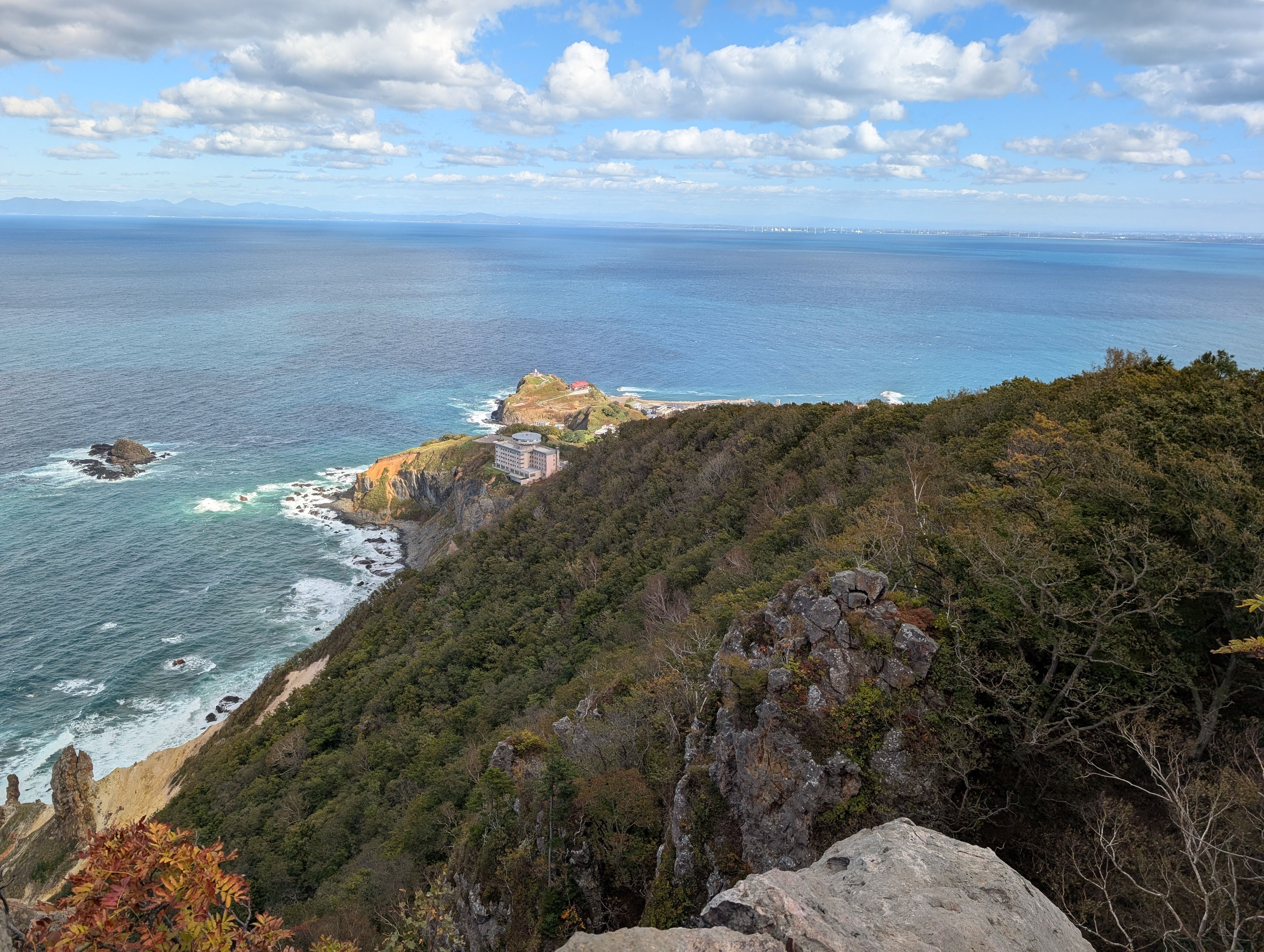 A view taken of the coastline from the top of a rocky cliff. It is a sunny day and the sea reflects the blue sky. It is early autumn and the leaves are beginning to change colour.